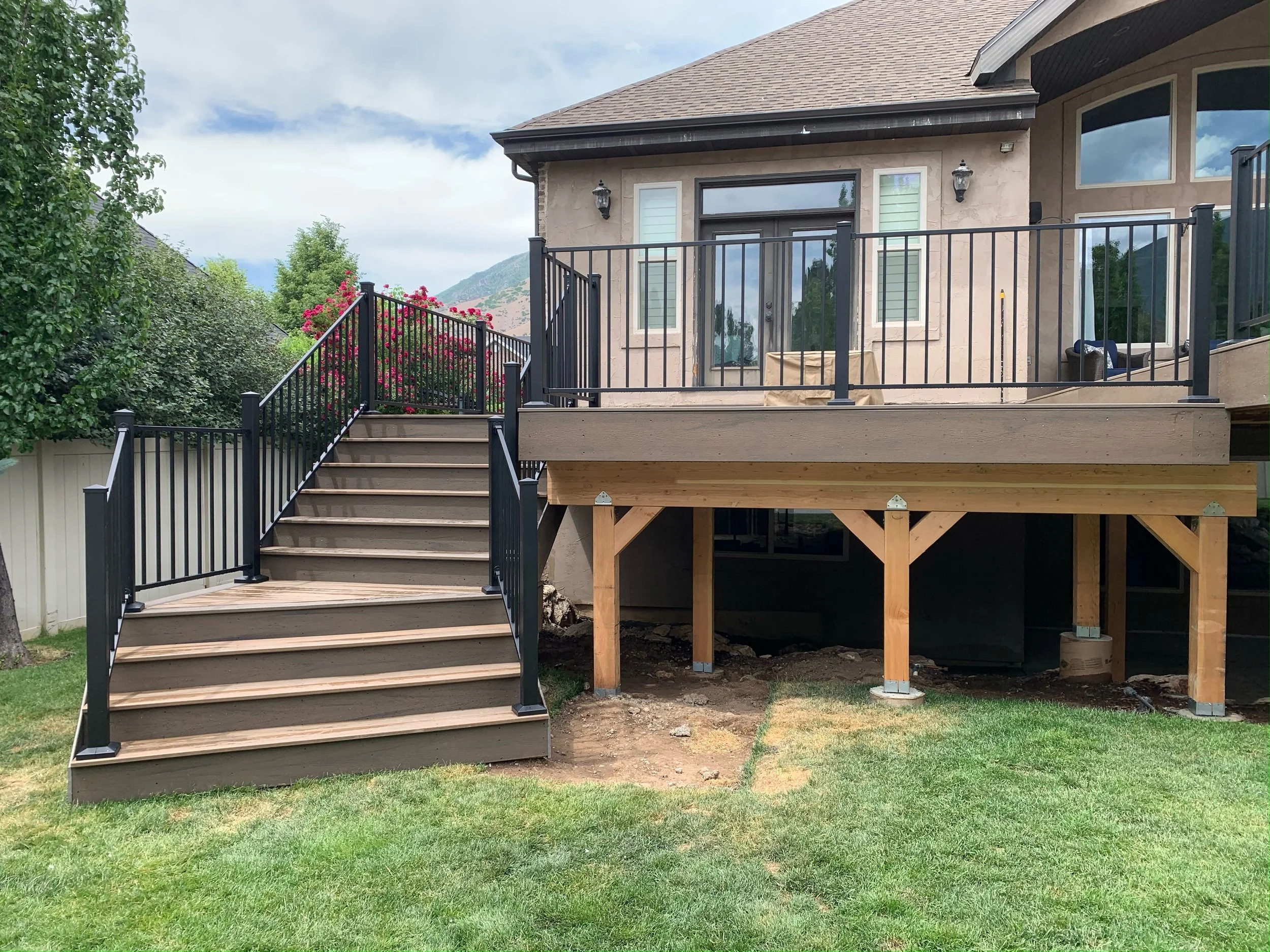 Wooden deck with black metal railing and stairs attached to beige house with sliding glass door, outdoor furniture, and mountain scenery in the background.