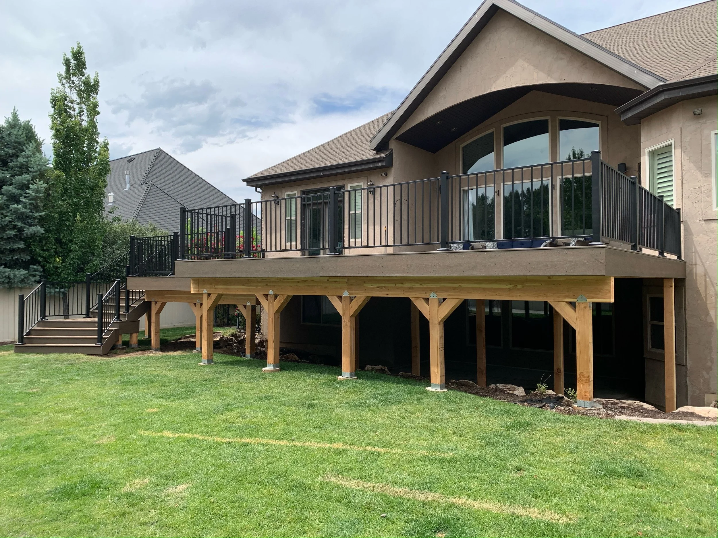 New wooden deck with black metal railings installed on the back of a house, with stairs leading down to a green lawn and neighboring houses visible in the background.