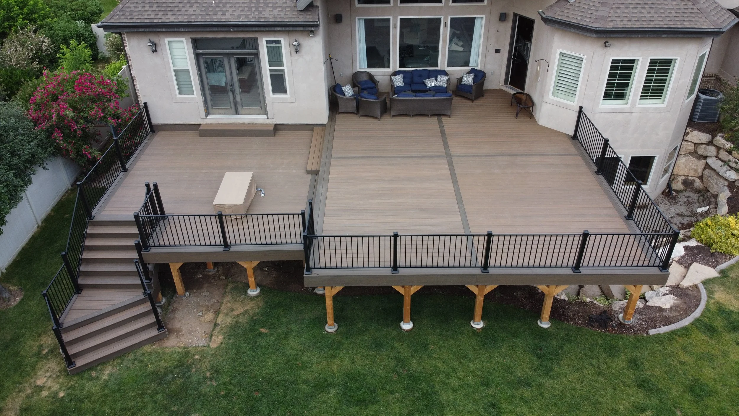 An aerial view of a large backyard deck attached to a house, with staircases on the left side, outdoor seating with blue cushions, and landscaped garden areas with rocks and plants.