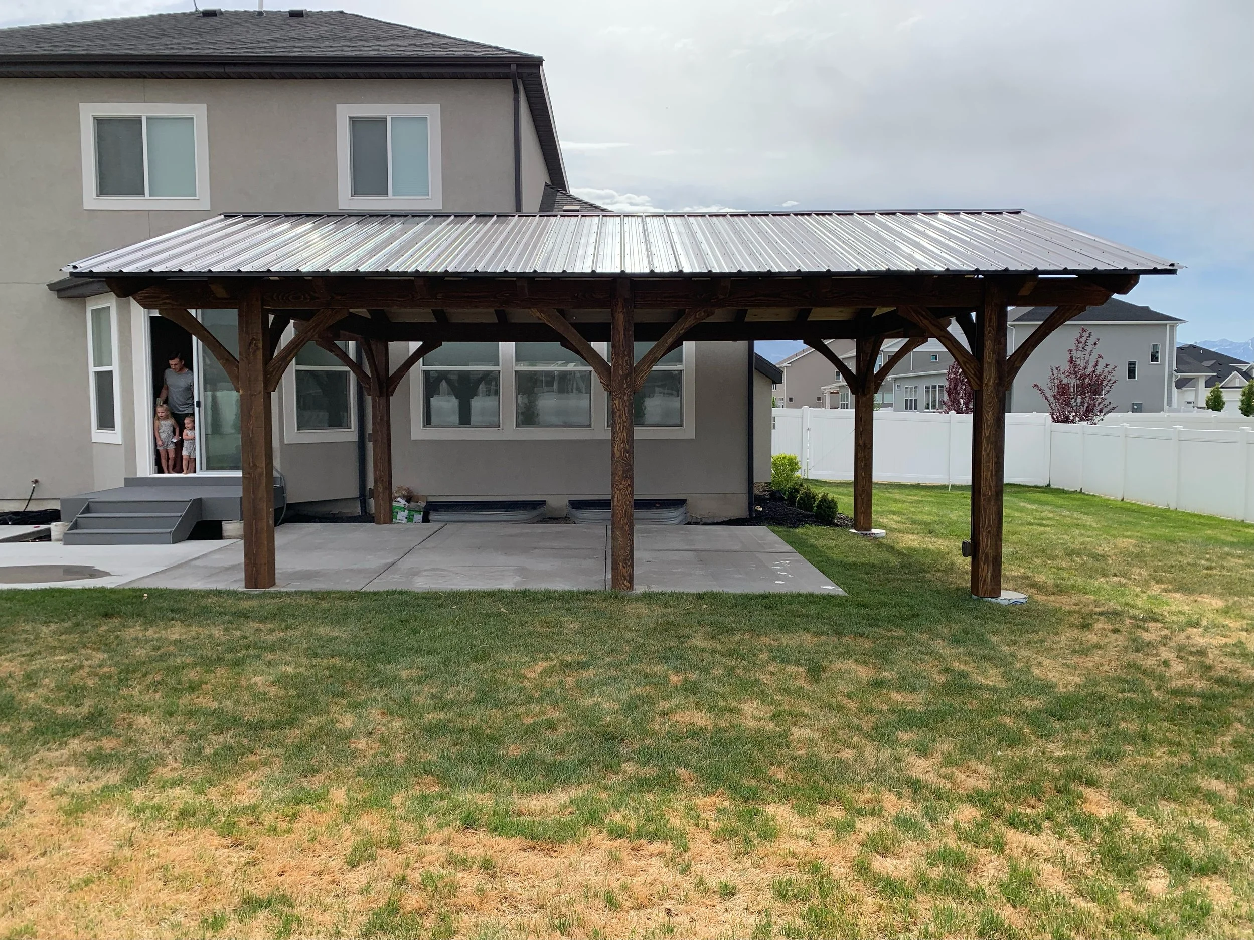 Backyard with newly built wooden covered patio with metal roof, concrete floor, and a house with two children and an adult standing in the doorway.