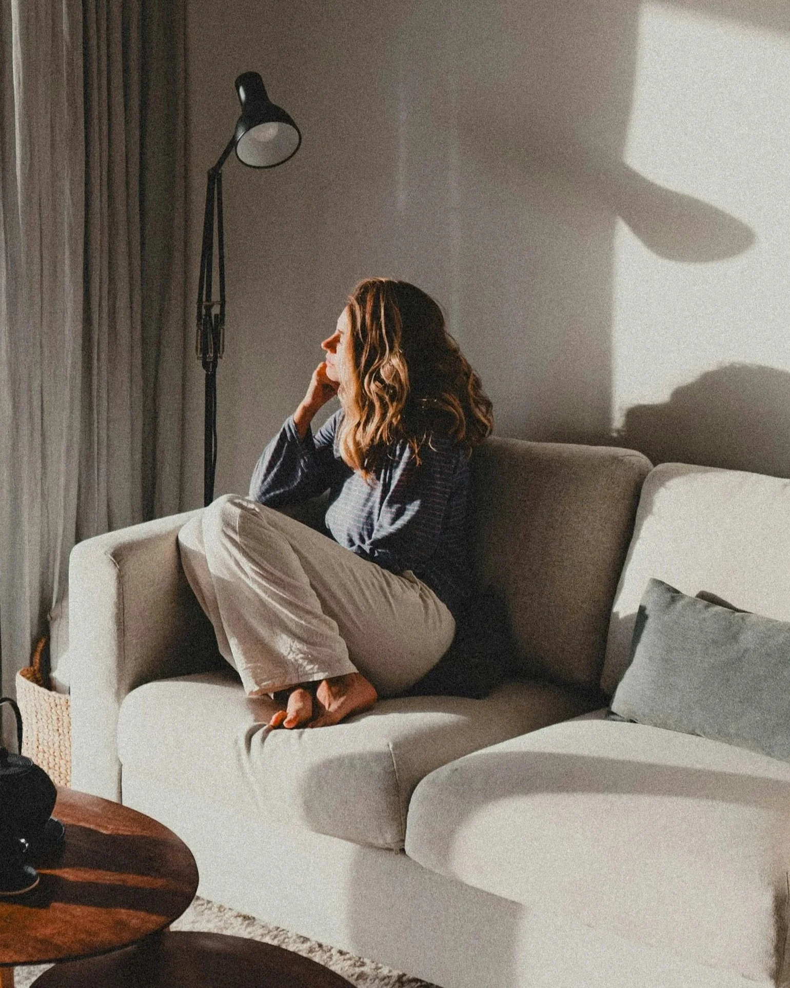 Adult sitting thoughtfully on a couch in soft natural light, representing trauma and emotional regulation support.