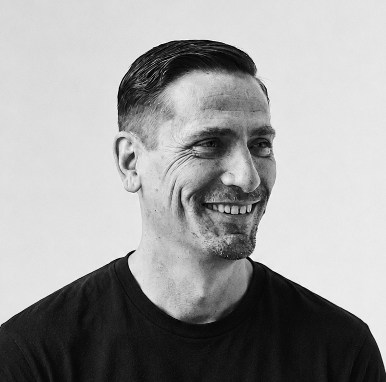 Black and white portrait of a smiling man looking to the side, with short dark hair slicked back, wearing a black t-shirt, against a plain light background.
