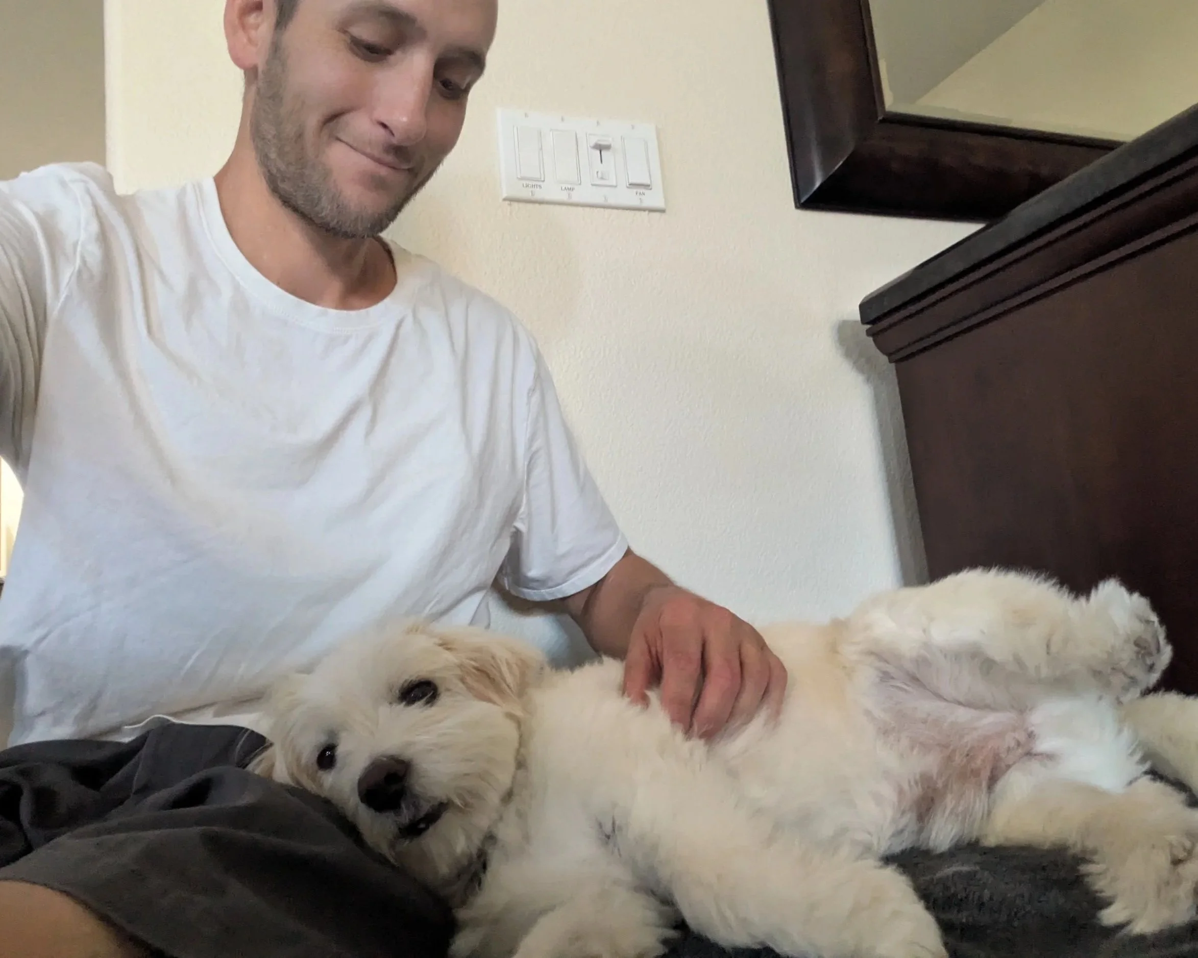 A man in a white t-shirt smiling while lying on a bed with a large, fluffy white dog on its back, petting the dog.