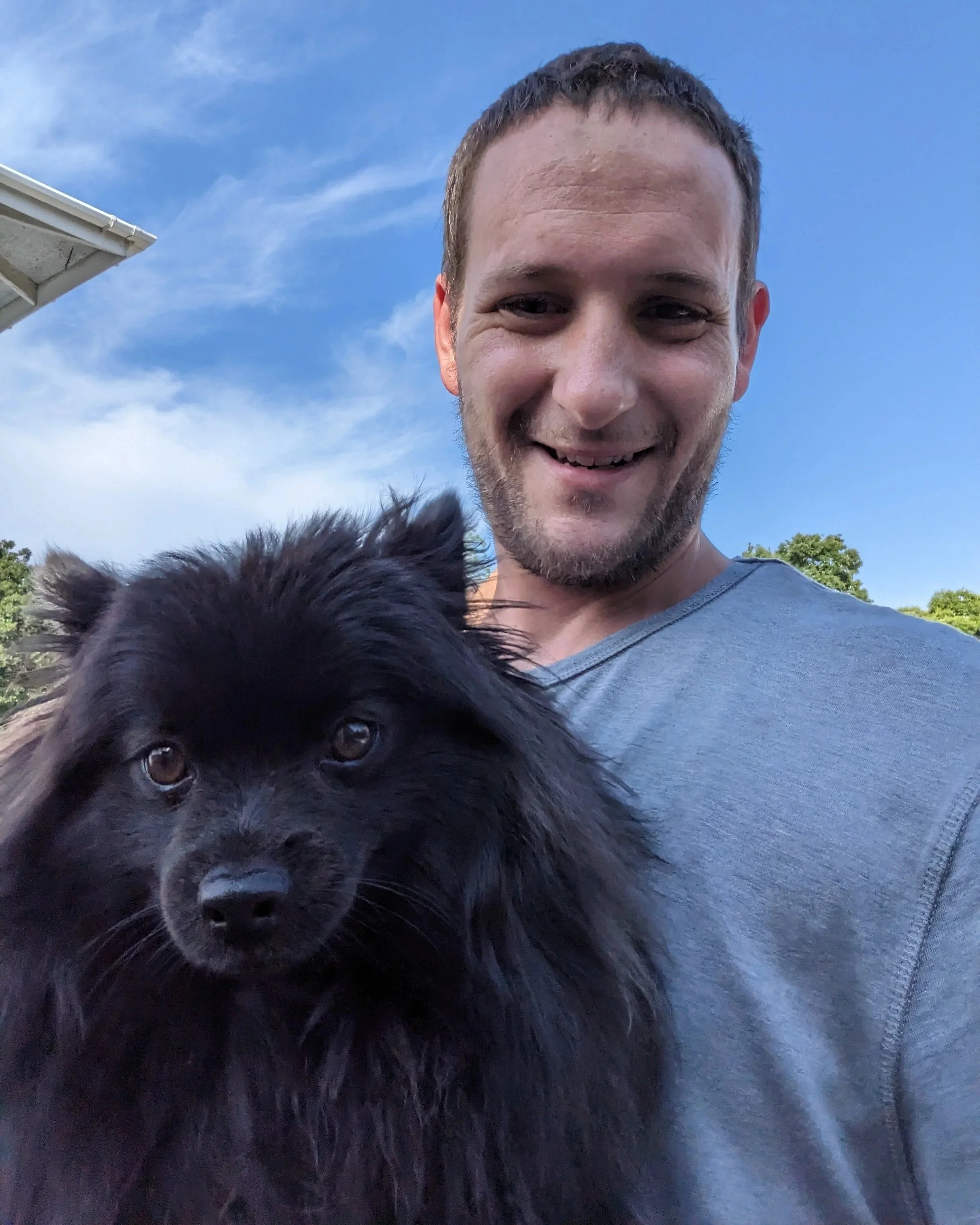 A man smiling outdoors holding a black dog with thick fur against a blue sky with some clouds and greenery in the background.