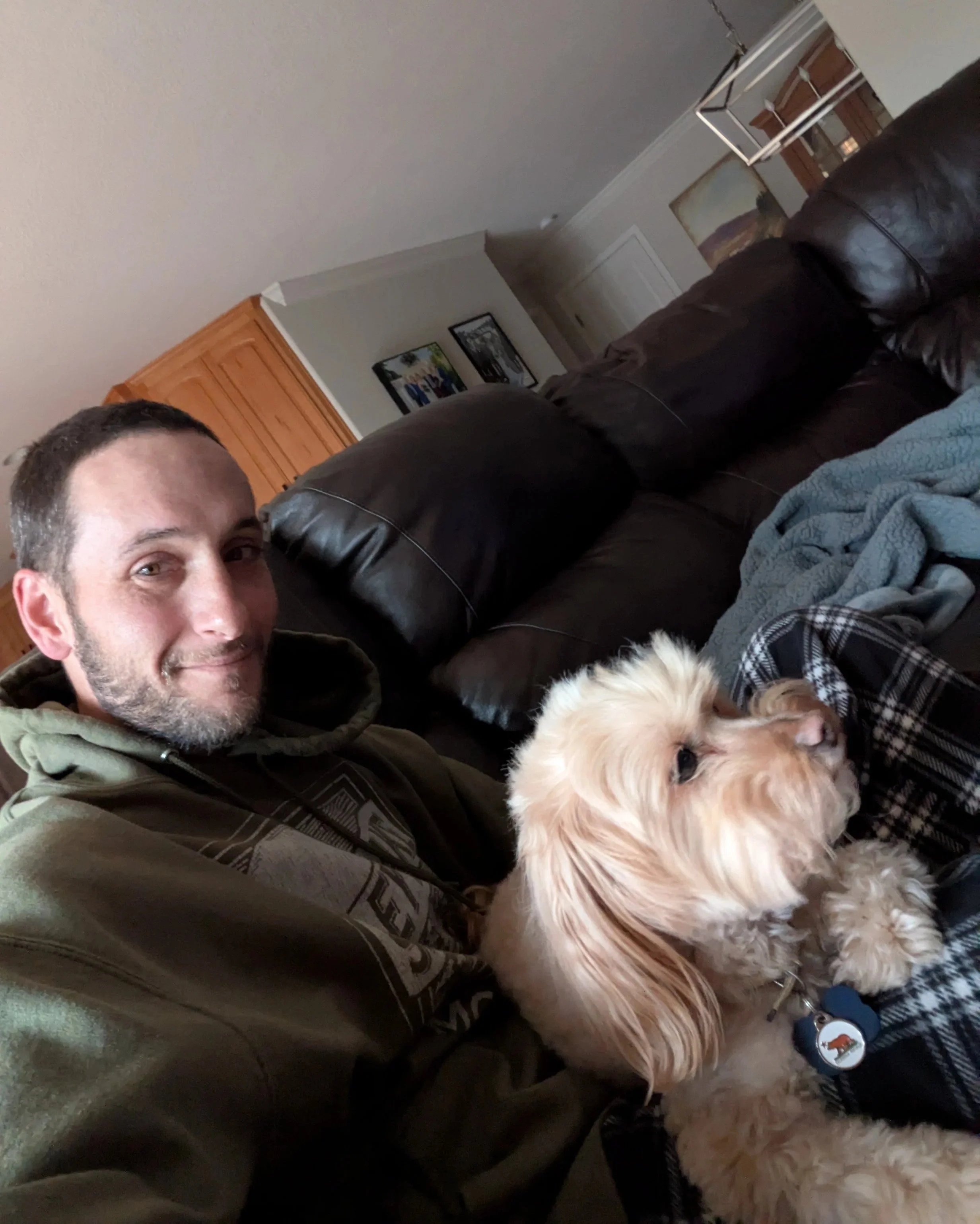 A man sitting on a brown leather couch with a light-colored dog, both looking to the right, in a living room with framed pictures on the wall.