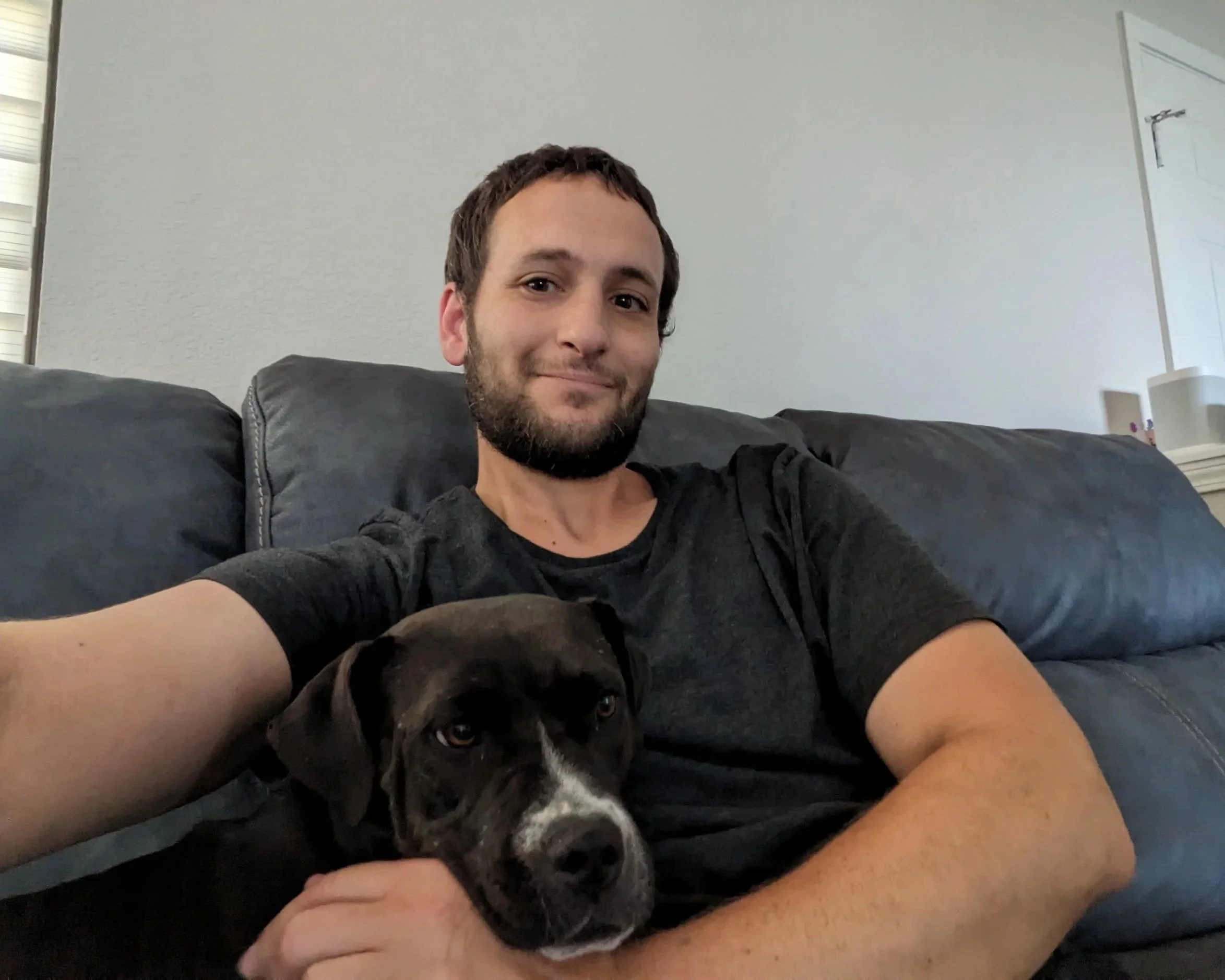 A man sitting on a black leather couch holding a black and white dog with a white stripe on its nose, in a room with white walls and a closed white door.