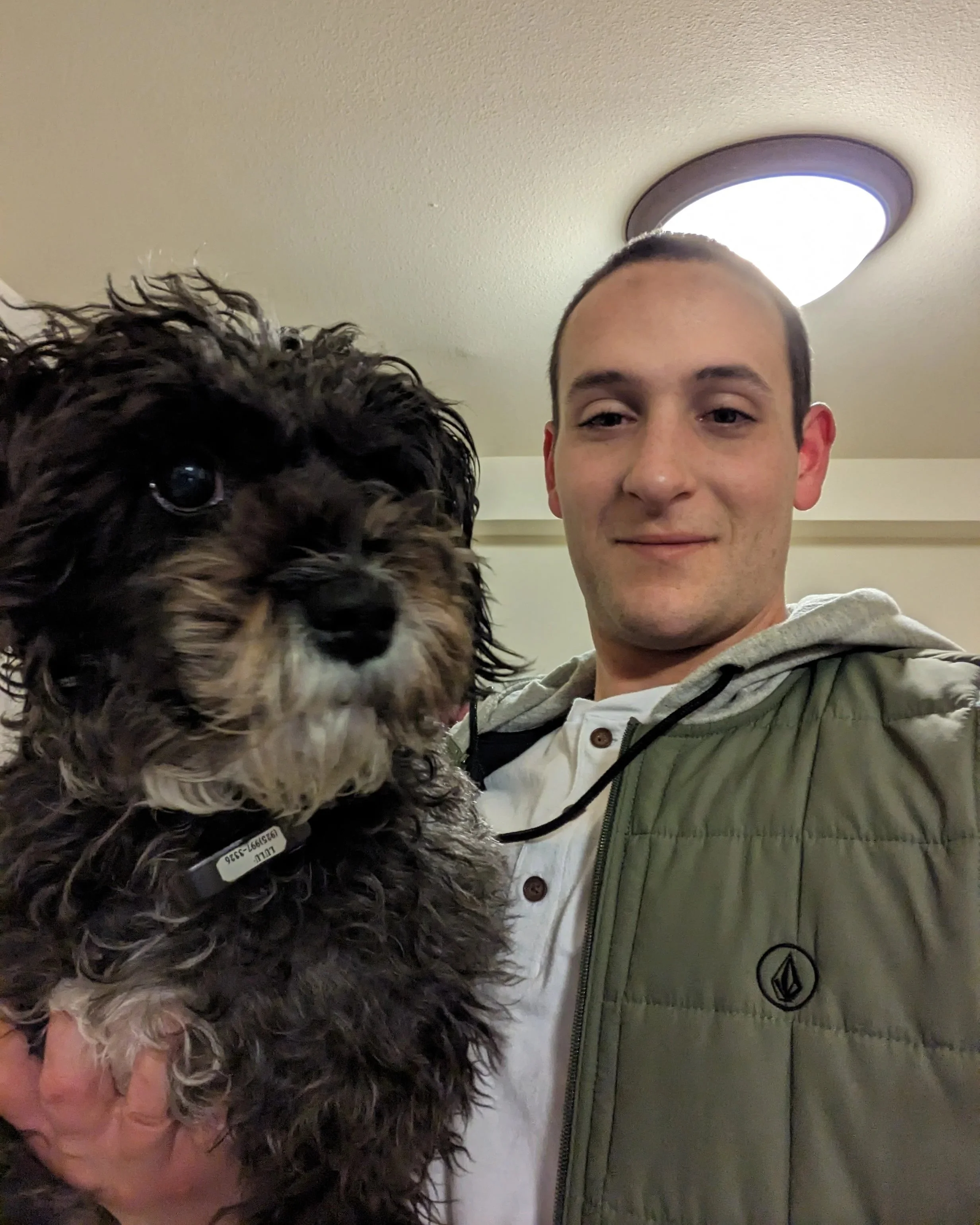 A young man with short hair and a slight smile, wearing a white shirt and green jacket, holding a small black and gray dog with curly fur inside a room with a ceiling light.