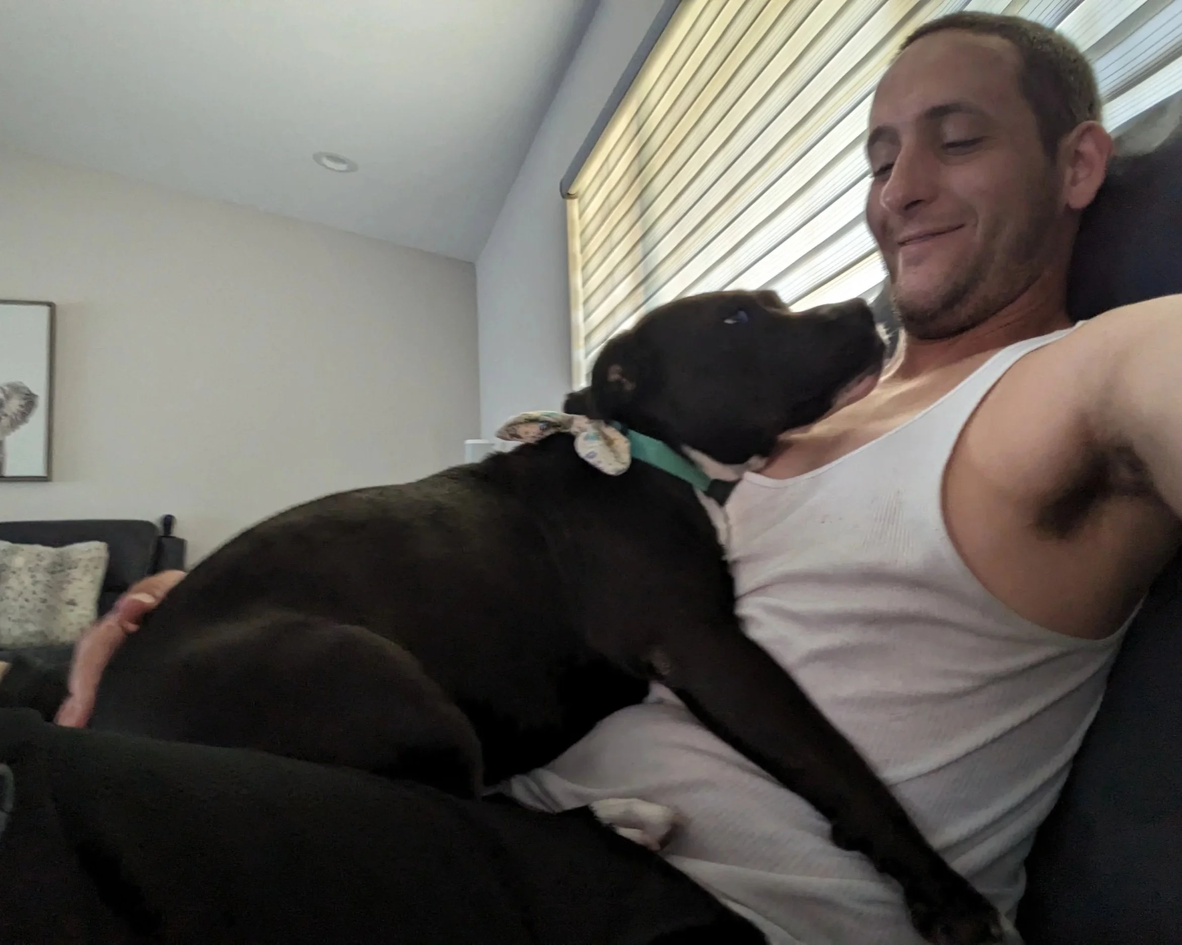 A man lying on a couch with a black puppy on his chest, smiling as the puppy nuzzles him.