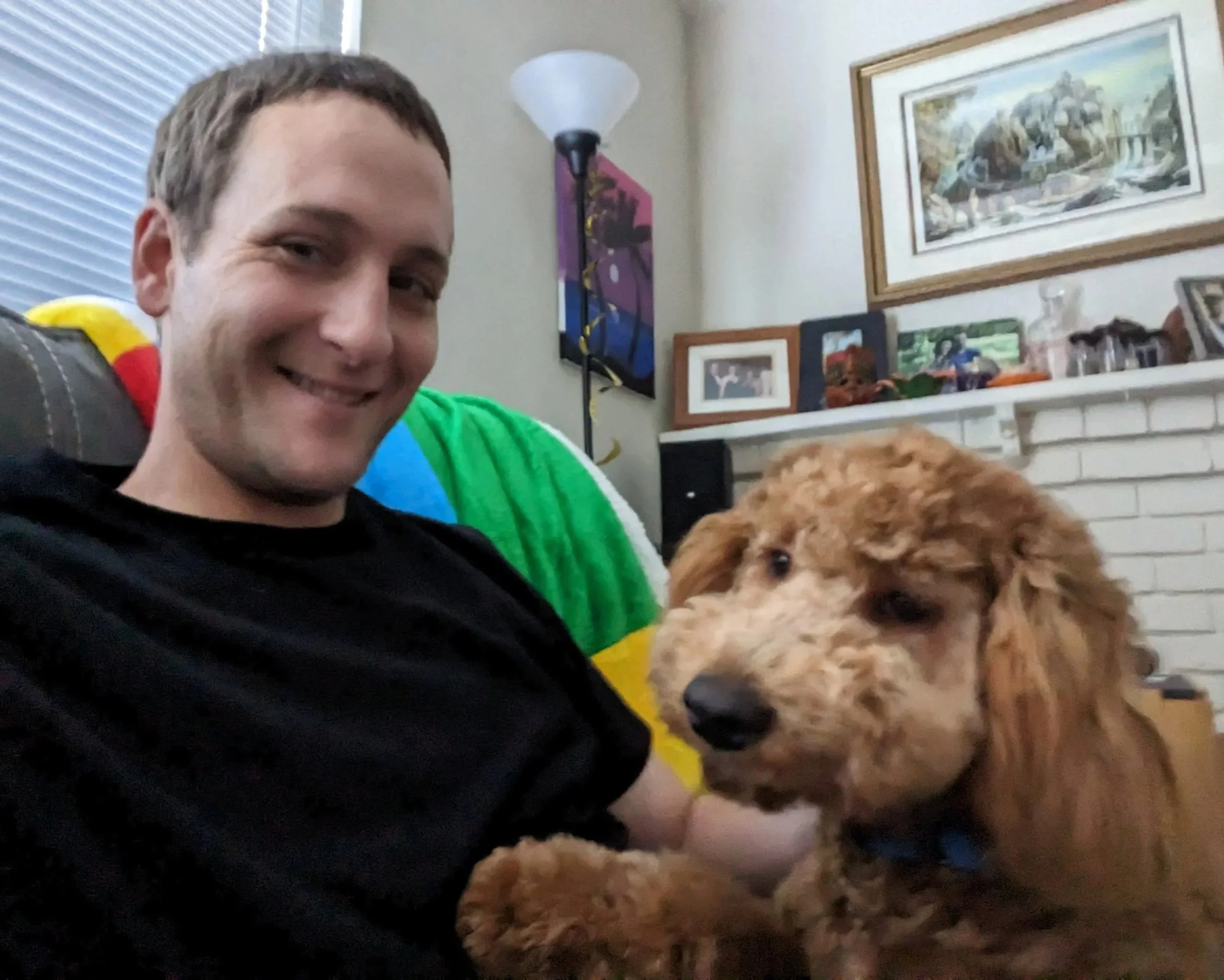 A smiling man sitting on a couch with a brown, curly-haired dog next to him in a living room.