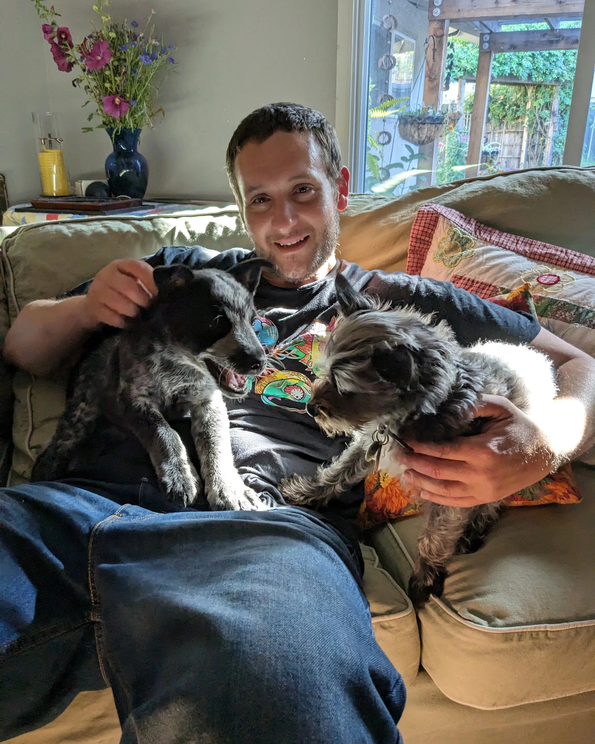 A man sitting on a sofa holding two puppies in his lap, smiling at the camera.