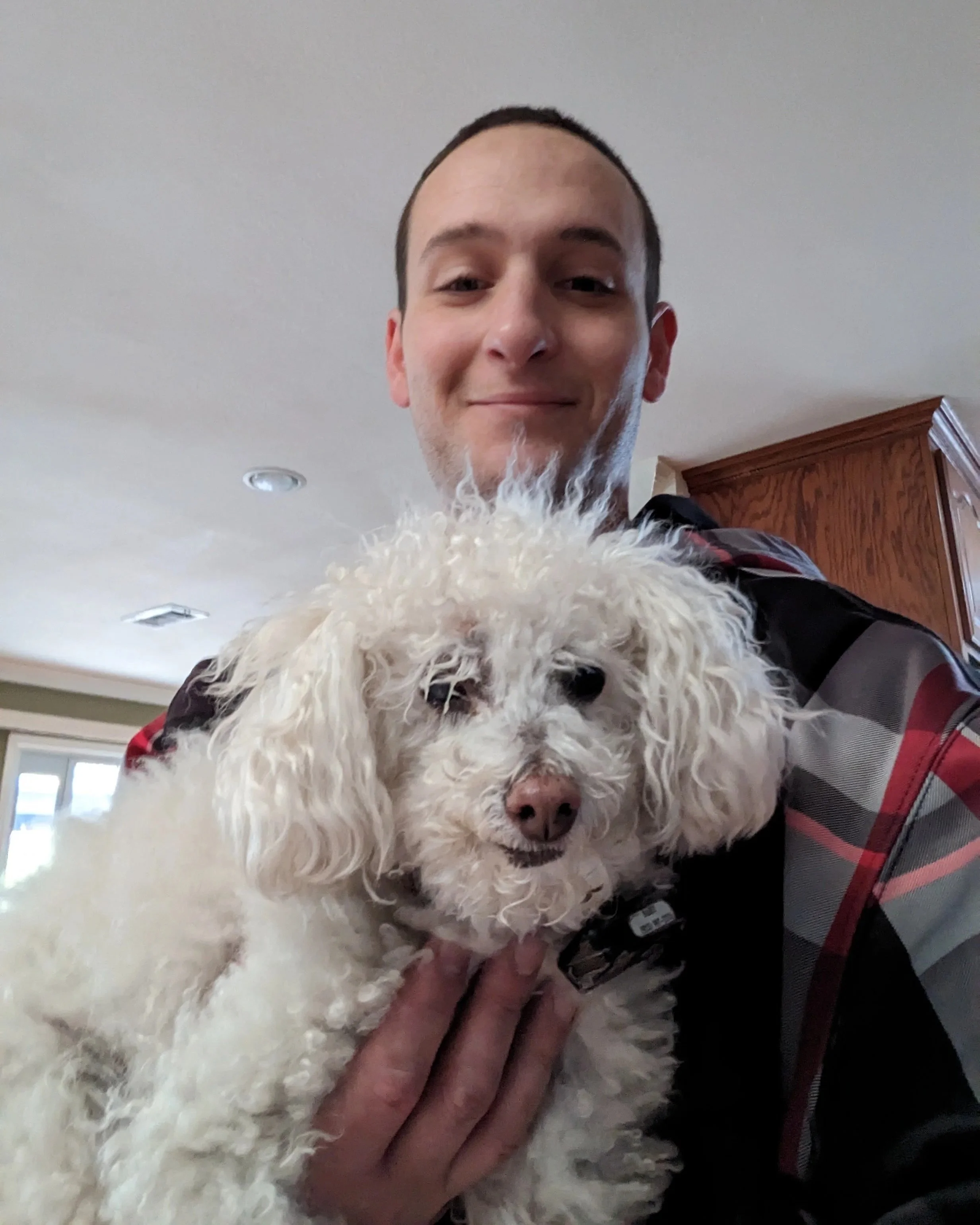 A young man smiling while holding a small, curly-haired white dog indoors.
