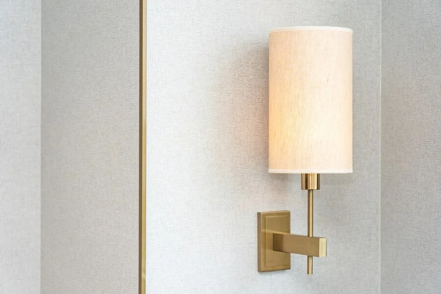 Wall-mounted lamp with a brass arm and beige shade in a room with light-colored textured wallpaper.