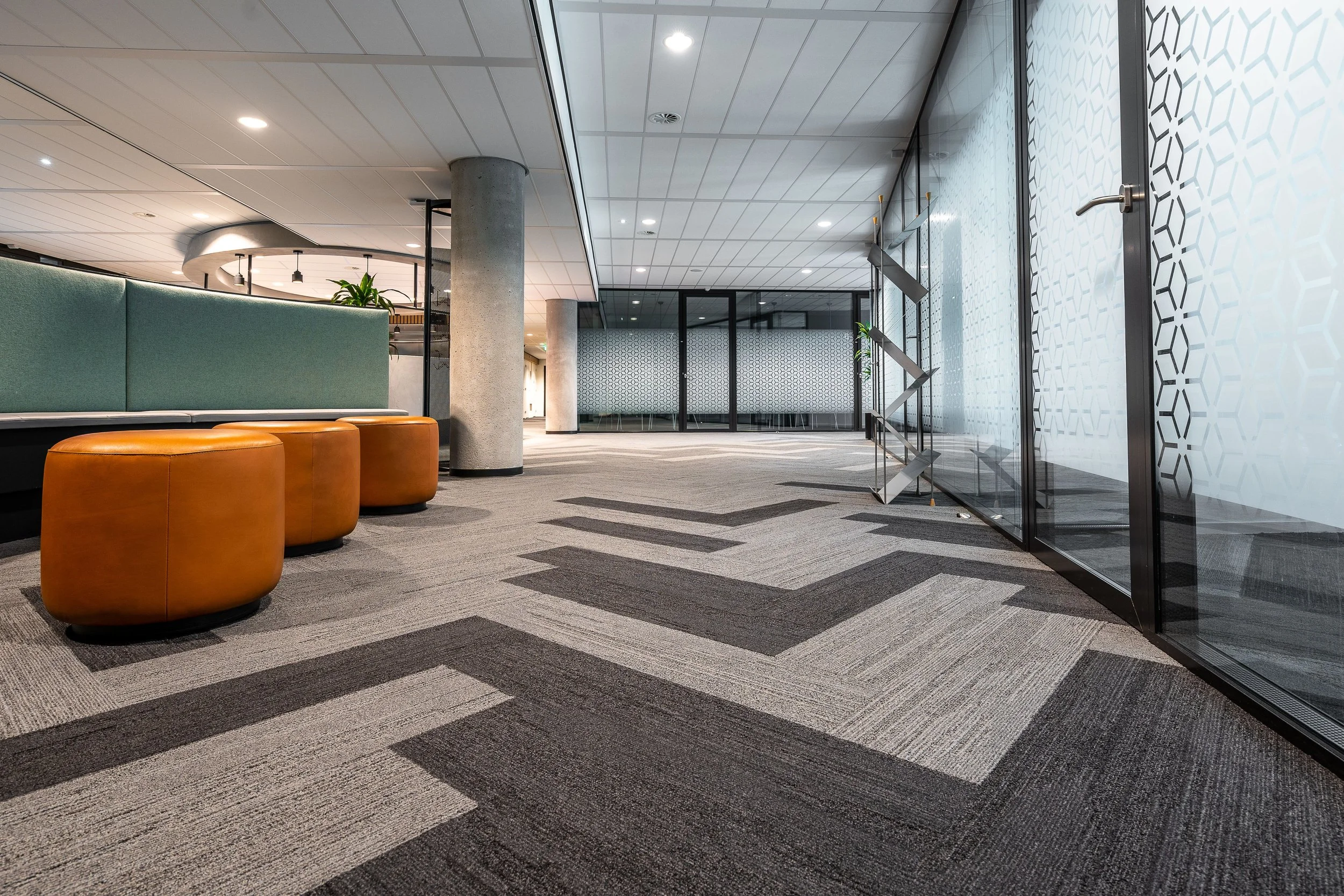 Modern office lobby with patterned carpet, orange ottomans, green seating area, glass walls, and decorative elements.