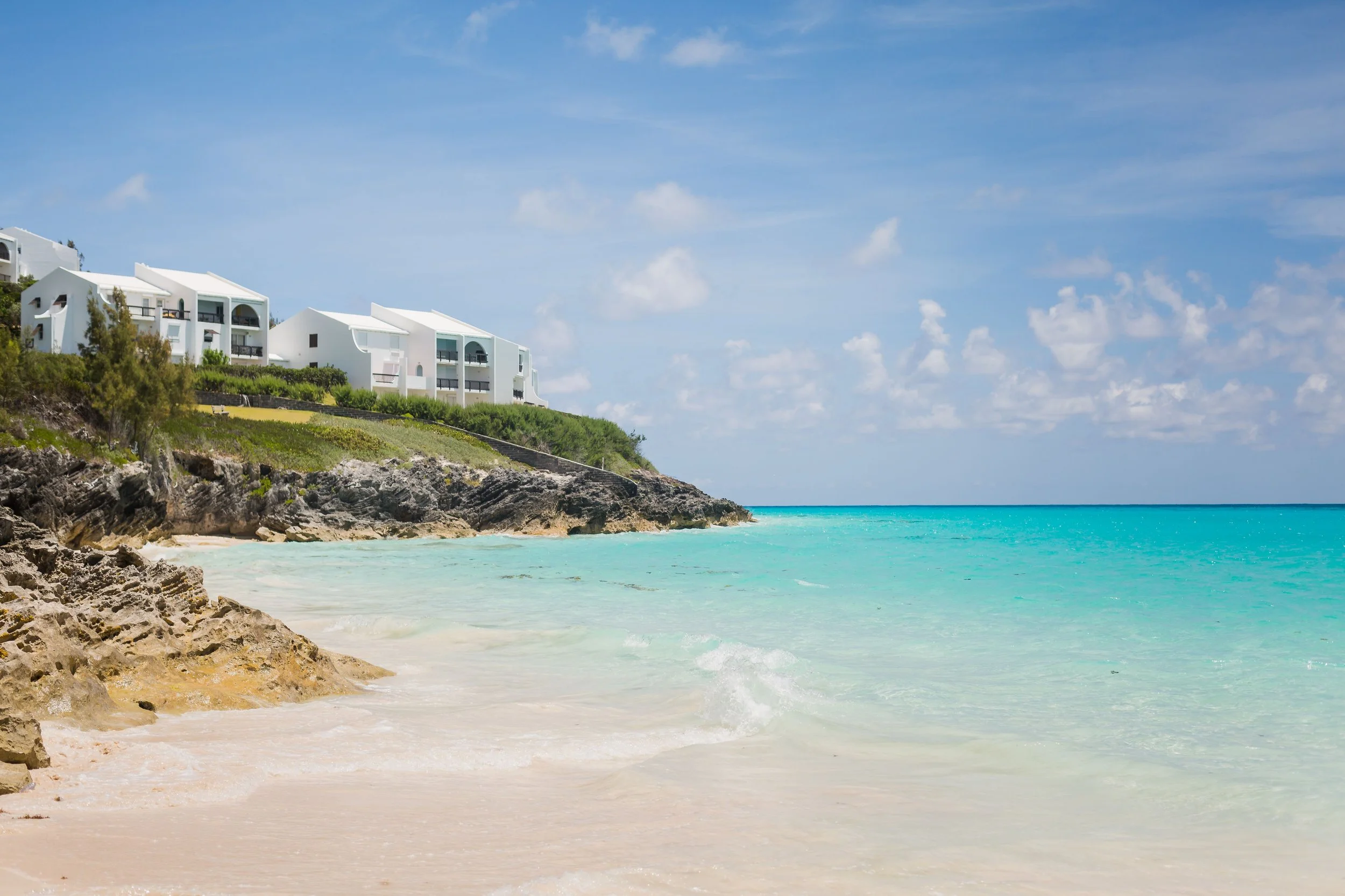 White buildings with dark balconies on a green hill overlooking a beach with rocky shoreline, turquoise water, and blue sky with scattered clouds.