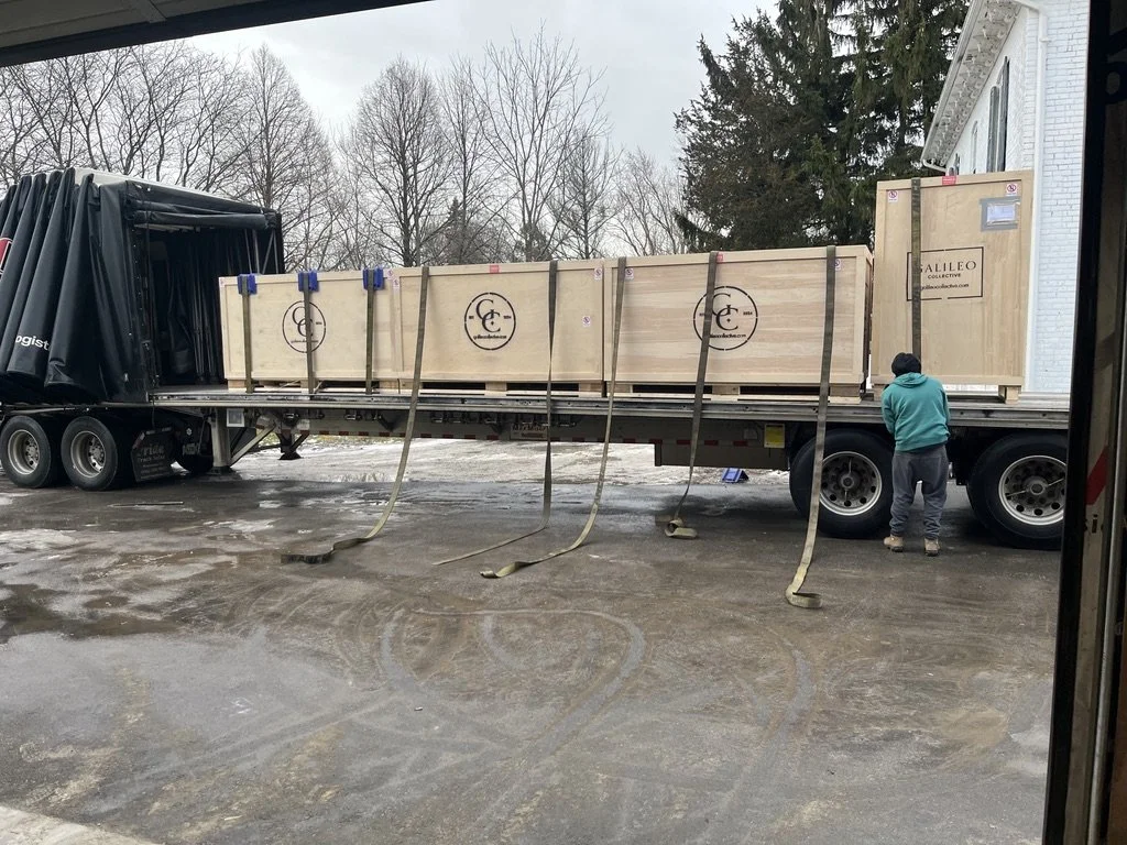 A person in a teal hoodie and gray sweatpants working on a loaded flatbed truck with large wooden crates in a driveway, with leafless trees in the background.