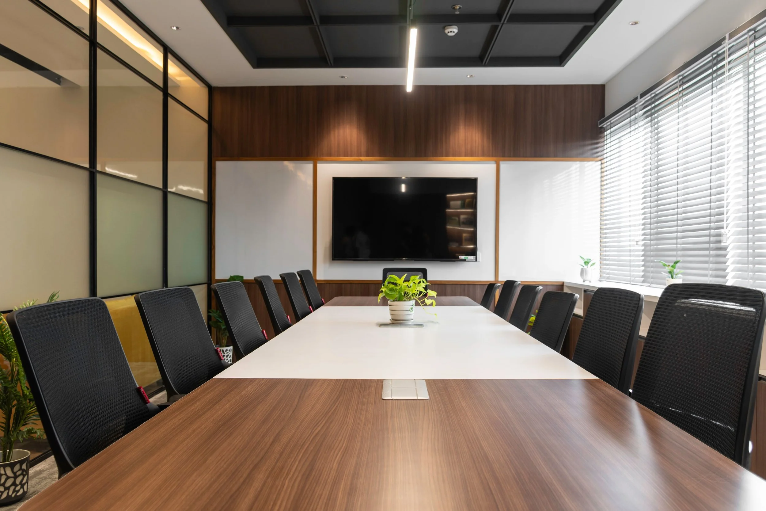A modern conference room with a long wooden table surrounded by black chairs, a large wall-mounted TV, and large windows with blinds letting in natural light, decorated with small potted plants.