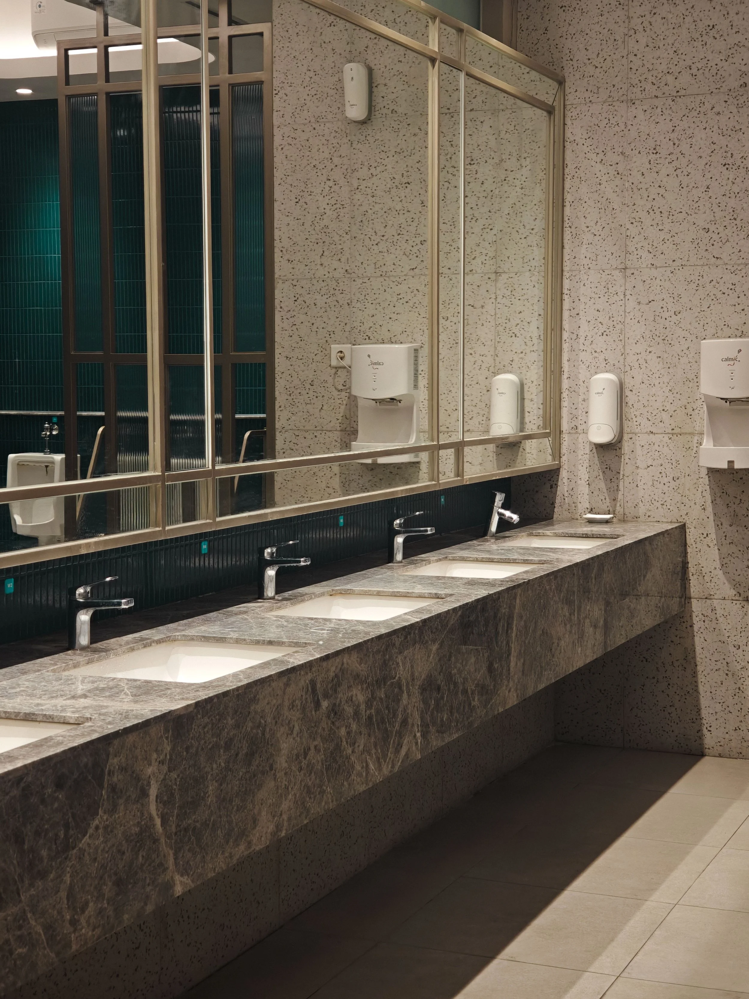 A public restroom with a long granite countertop featuring multiple sinks, a large mirror, and soap dispensers on the wall. The background includes a partition with teal tiled walls and urinals.