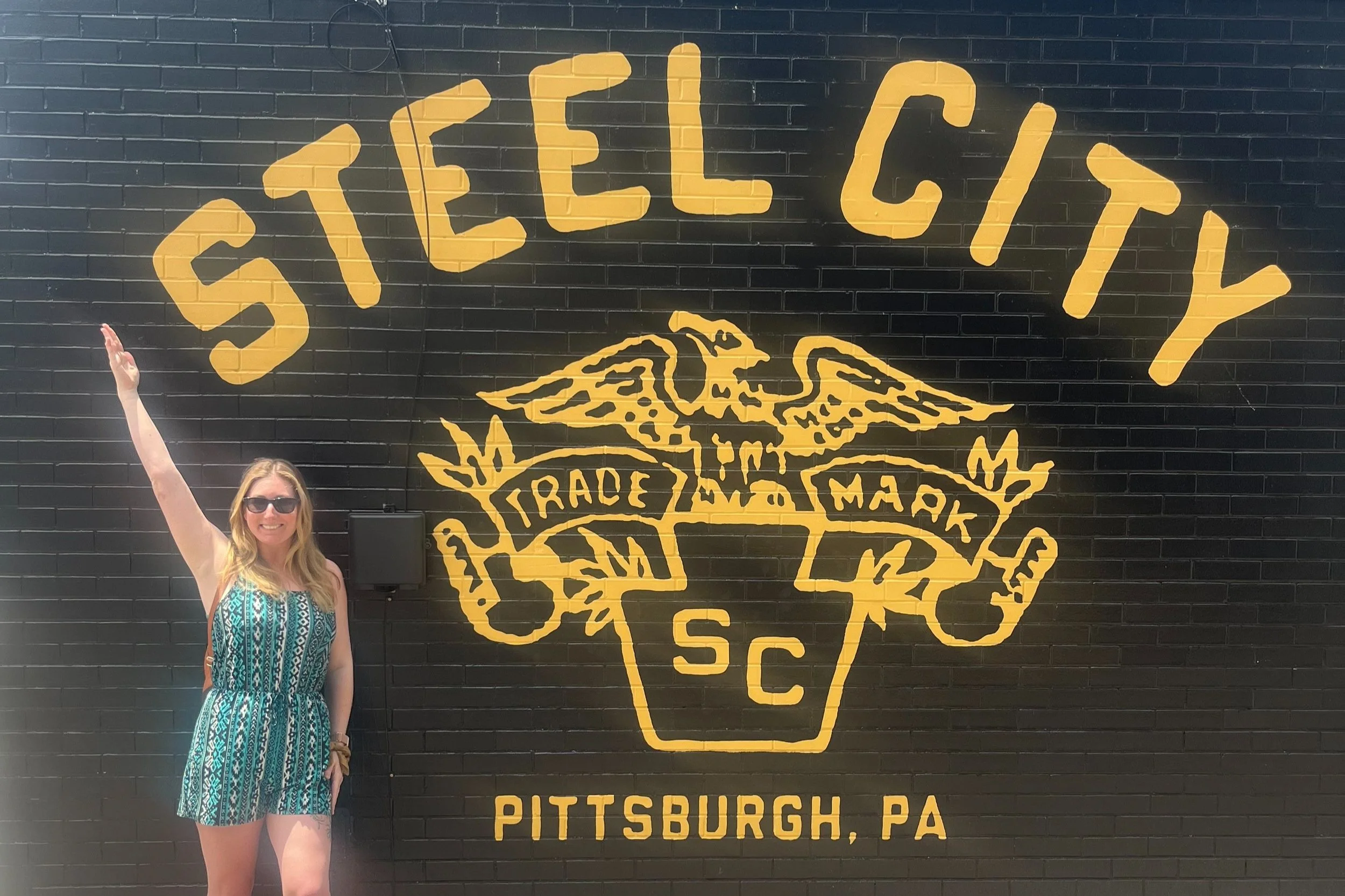 Woman in sunglasses and a patterned summer dress standing next to a large black wall with yellow graffiti-style logo of Steel City and Pittsburgh, PA.