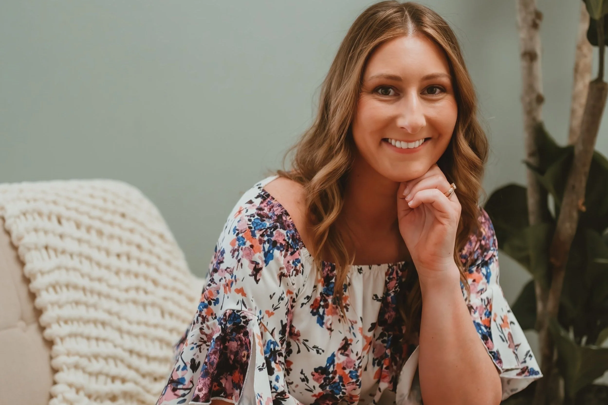 A woman with wavy brown hair smiling, resting her chin on her hand, sitting on a beige sofa with a knit throw blanket and a plant nearby, against a light green wall.