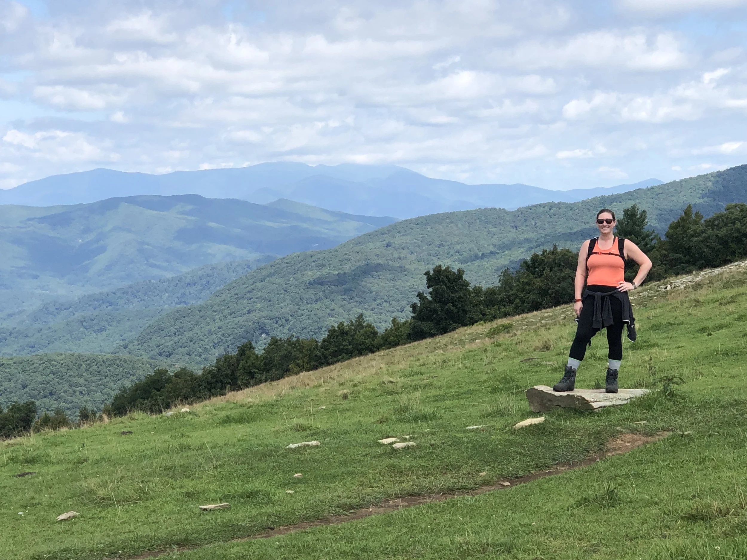 A woman standing on a large rock in a green field with rolling hills and mountains in the background, wearing hiking gear and sunglasses.