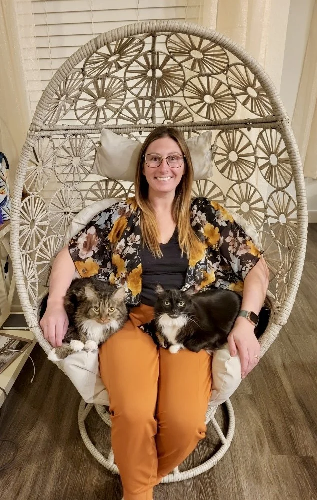 A woman with glasses sitting in a wicker hanging chair with a floral pattern, smiling, with two black and white cats on her lap.