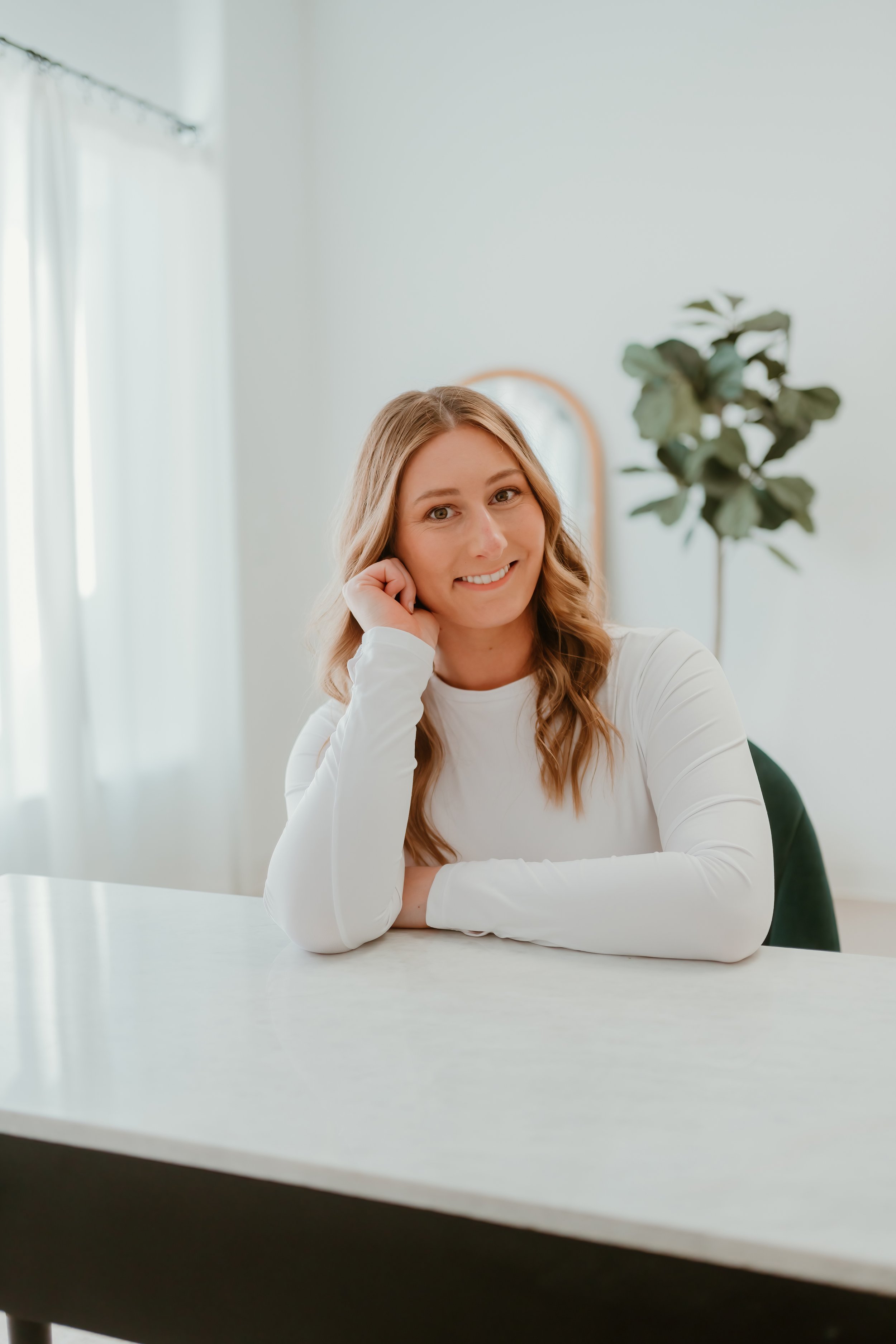 A young woman with wavy blonde hair, wearing a white long-sleeve shirt, sitting at a white table in a bright room with white curtains and a large green plant in the background, smiling at the camera.