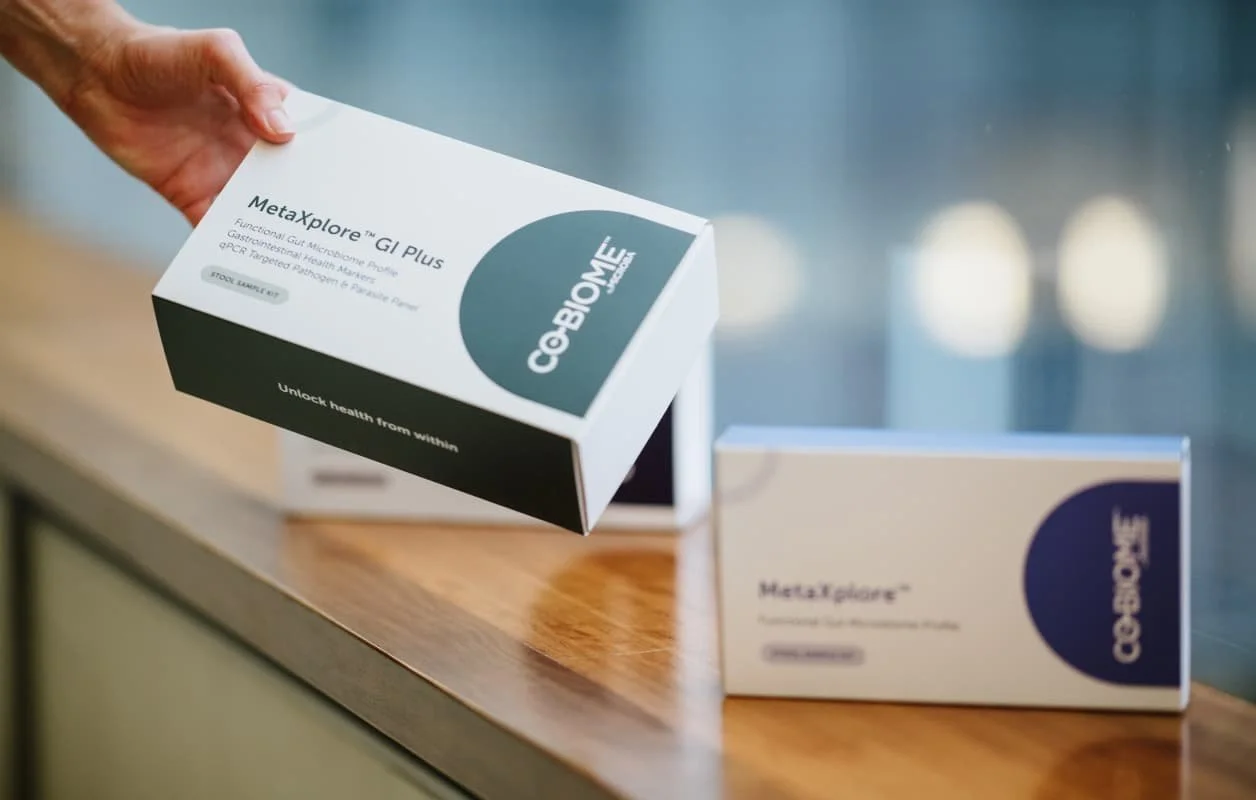 A person holding a box labeled 'MetaXplore GI Plus' among other similar boxes on a wooden surface.