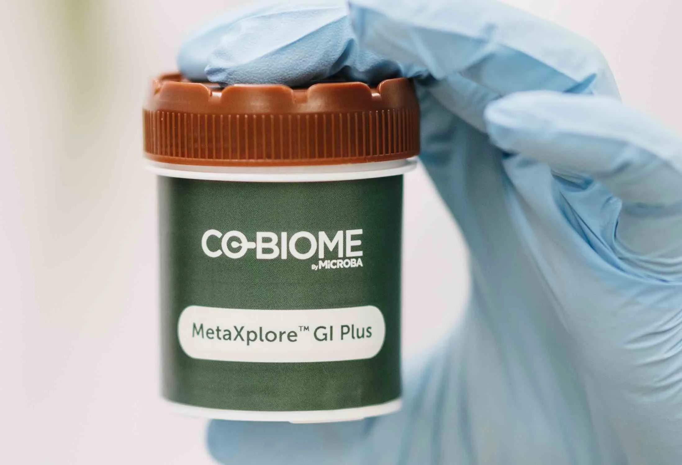 A gloved hand holding a jar labeled 'CO-BIOME by Microba' with the product name 'MetaXplore GI Plus' on a green label.