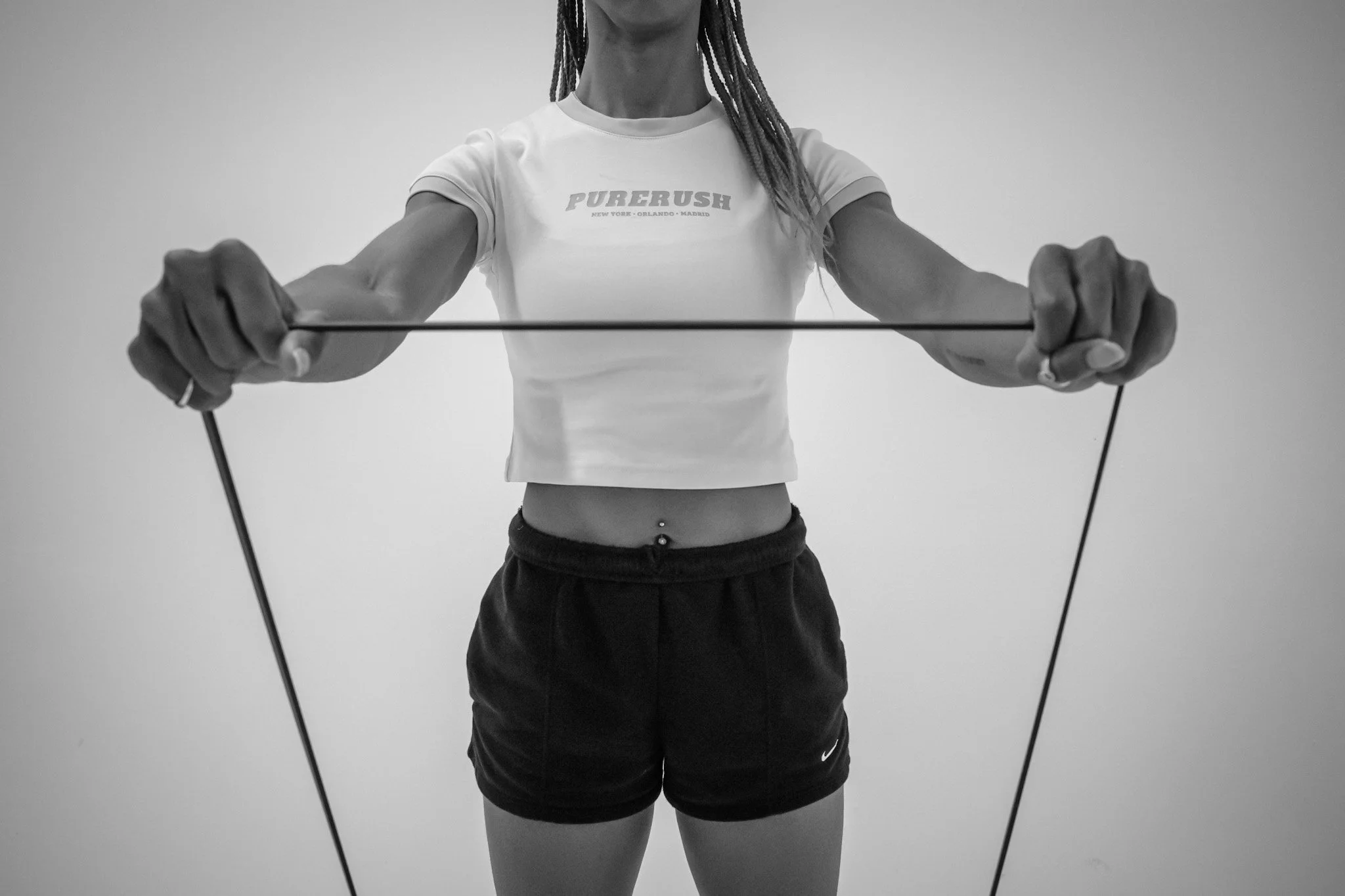 Person holding resistance band with both hands, wearing a white crop top and black shorts, standing against a plain background.