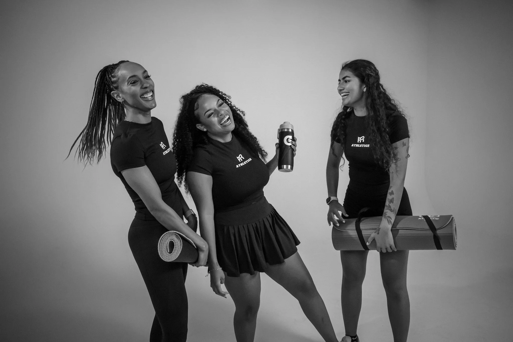 Three women wearing matching athletic t-shirts smiling and laughing while holding yoga mats and water bottles in a studio.