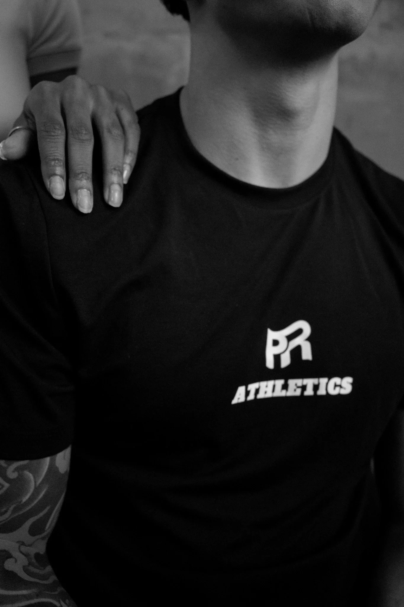 Close-up of a person wearing a black athletic t-shirt with the logo and text "PR Athletics." Another person is resting their hand on the shoulder of the person in the t-shirt.