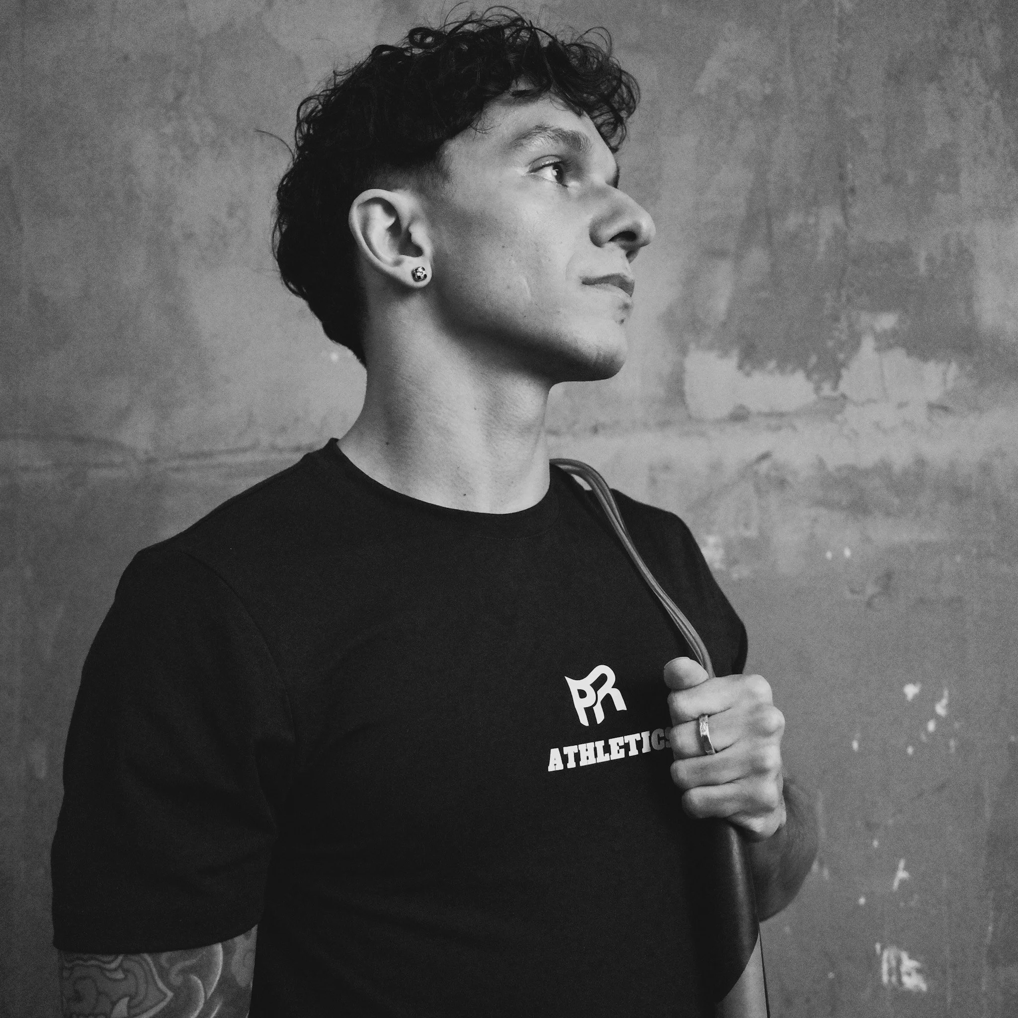 A young man with curly hair, earrings, and a tattoo on his arm, wearing a black athletic shirt, holding a jump rope over his shoulder, standing against a textured wall.