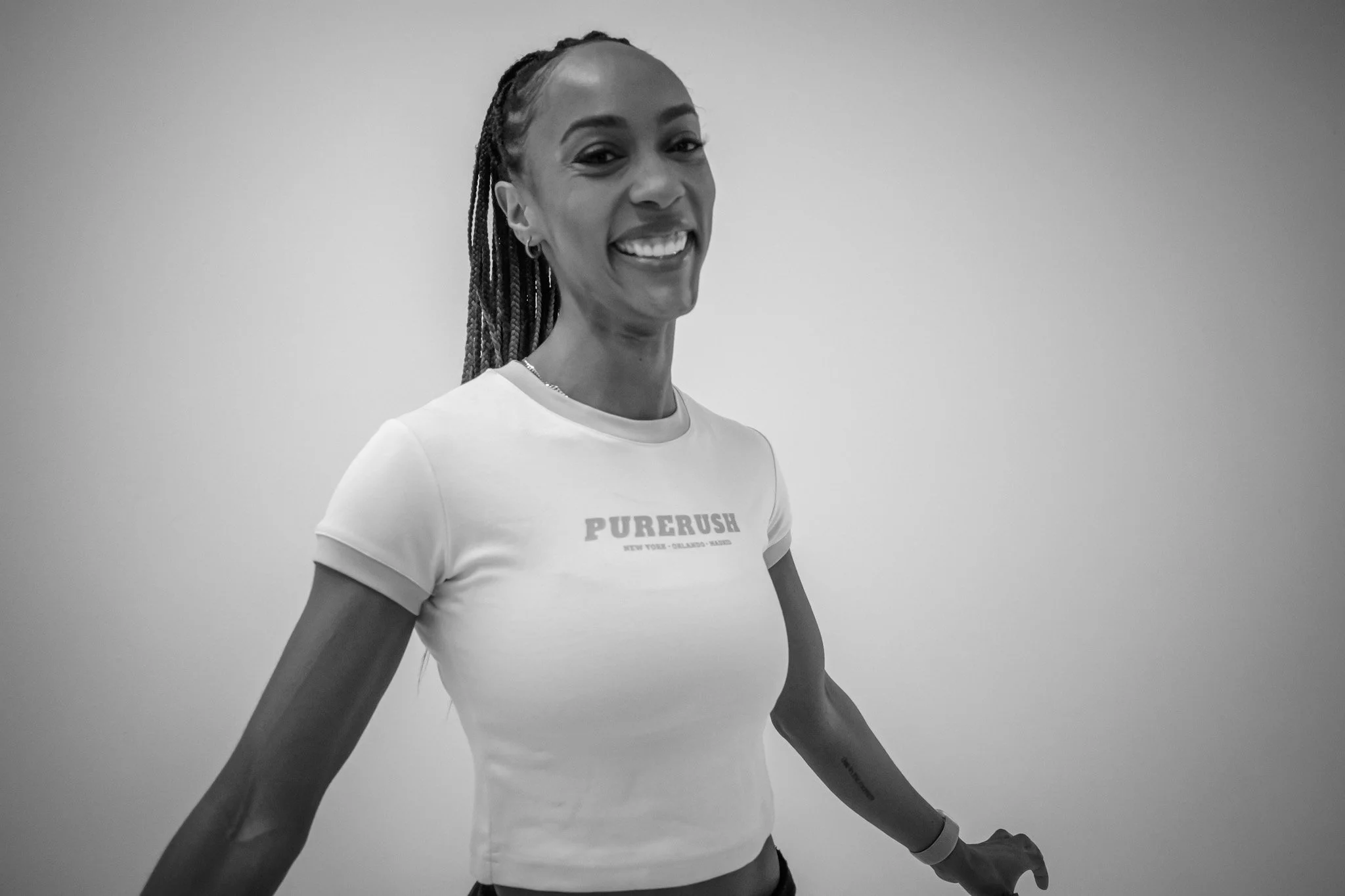 Smiling woman with braided hair wearing a white T-shirt with 'PURE RUSH' written on it, standing against a plain background.