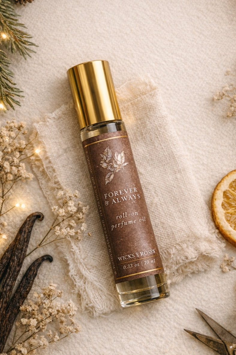 A bottle of Forever & Always roll-on perfume oil labeled Wicks & Roses, placed on a piece of fabric with dried flowers, vanilla pods, and a slice of dried orange surrounding it.