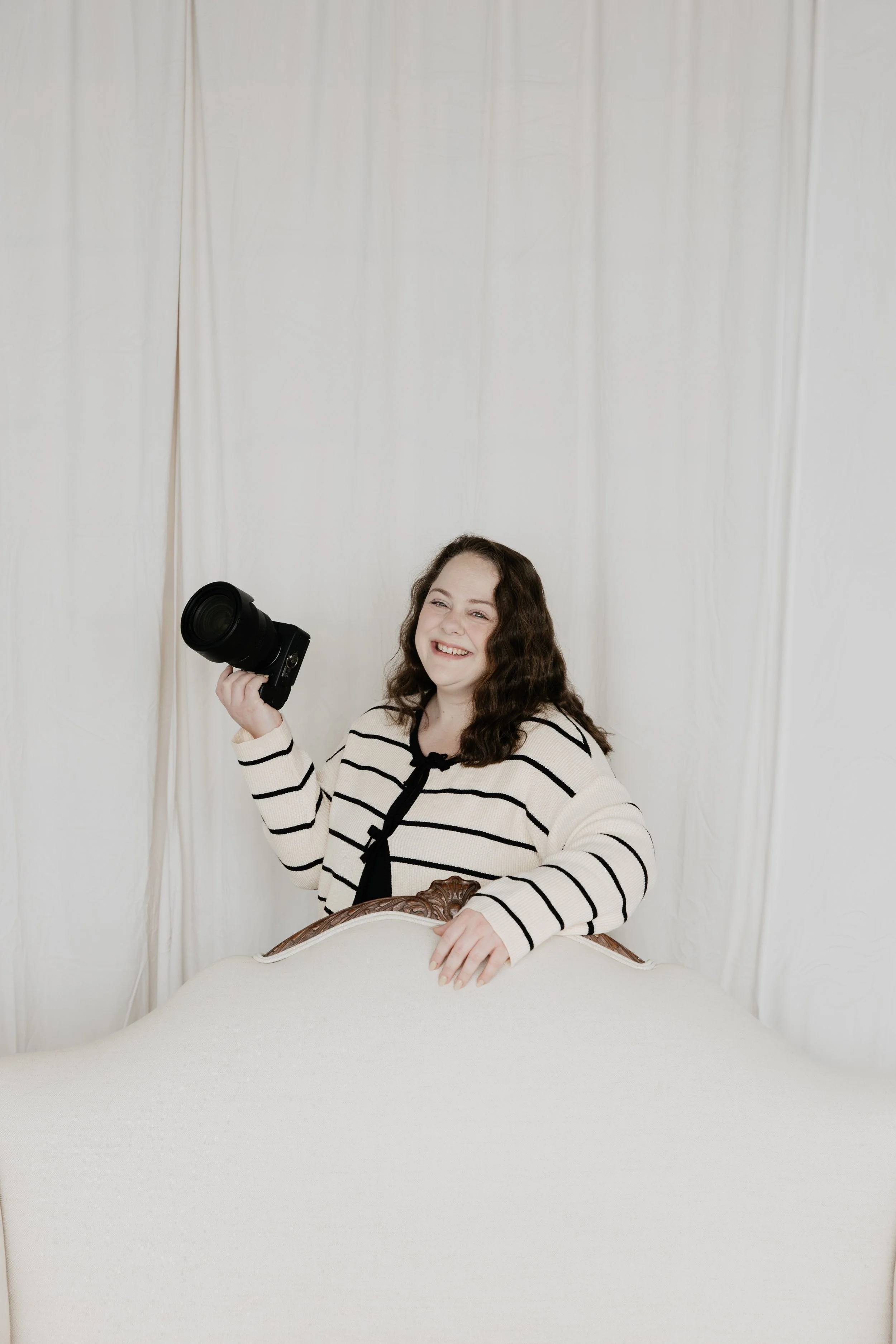 A smiling woman with brown wavy hair in a striped sweater holding a camera behind a white vintage-style sofa with a carved wooden back, against a white curtain background.