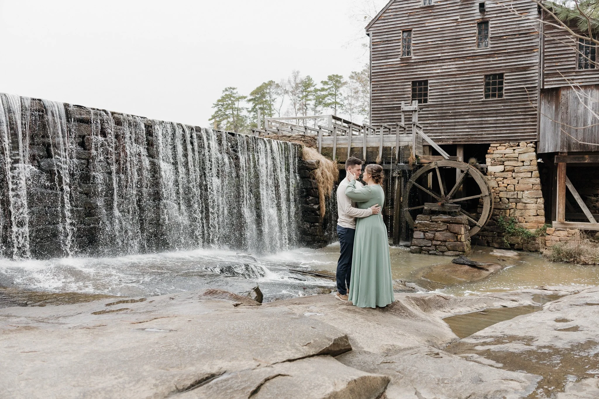 Engagement session at Yates Mill Historic Park in Raleigh, North CarolinaEngaged couple sharing a romantic moment beside the cascading waterfall and historic stone structure at Yates Mill.
