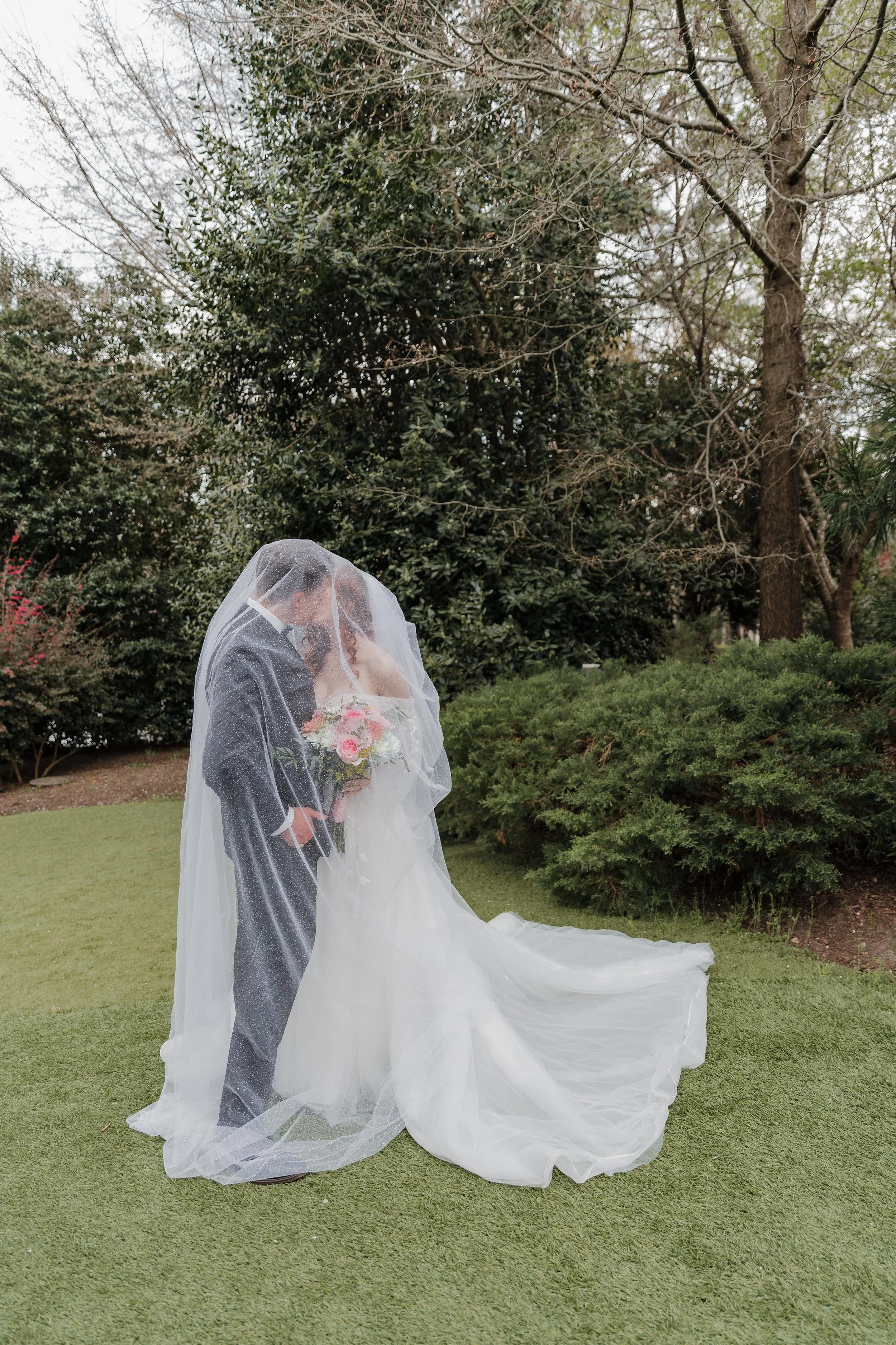 A bride and groom sharing a kiss under a wedding veil outdoors, with trees and greenery in the background.