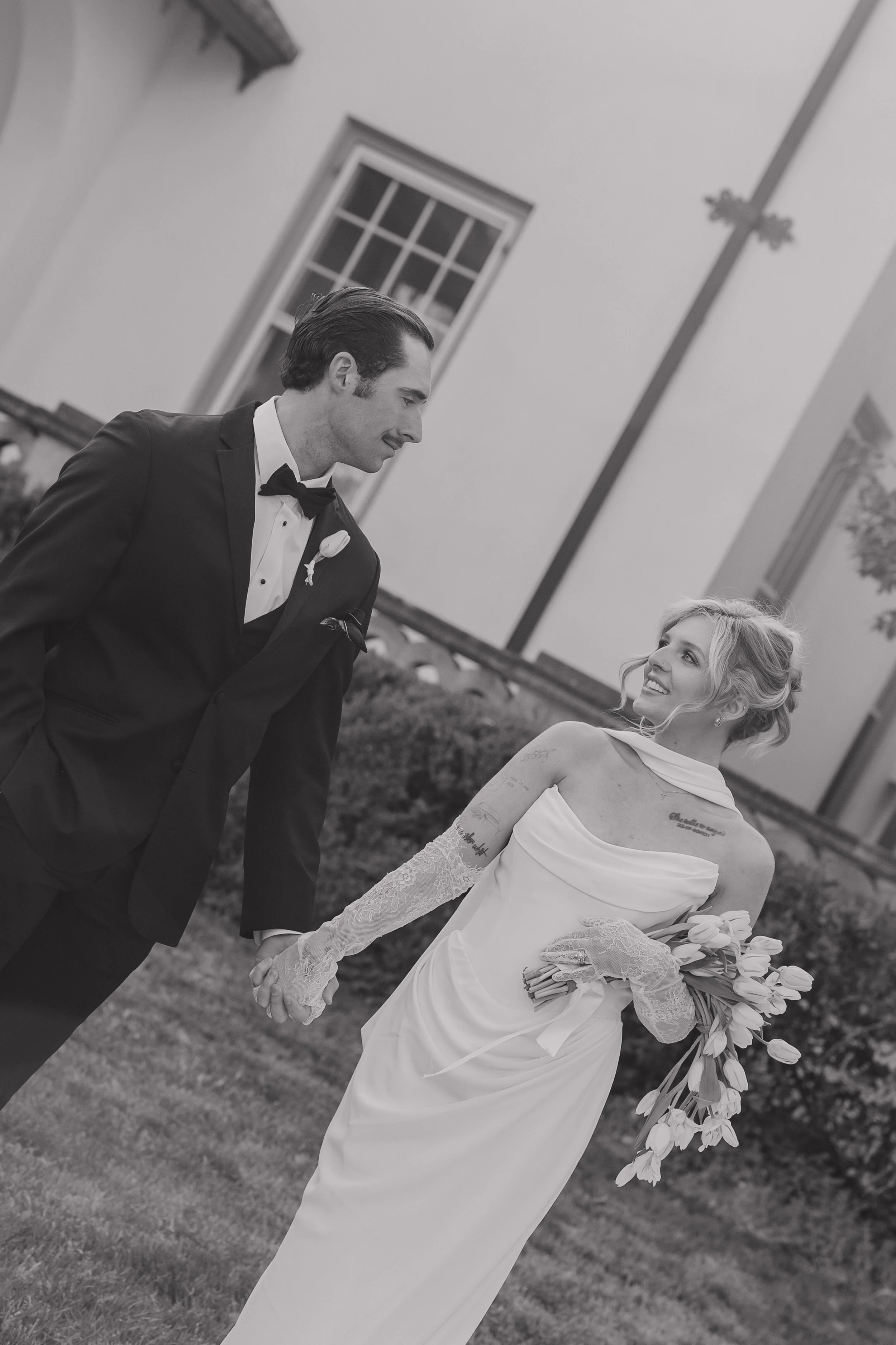 A black and white photo of a bride and groom holding hands and gazing at each other outdoors near a building with a window and a cross on the wall.