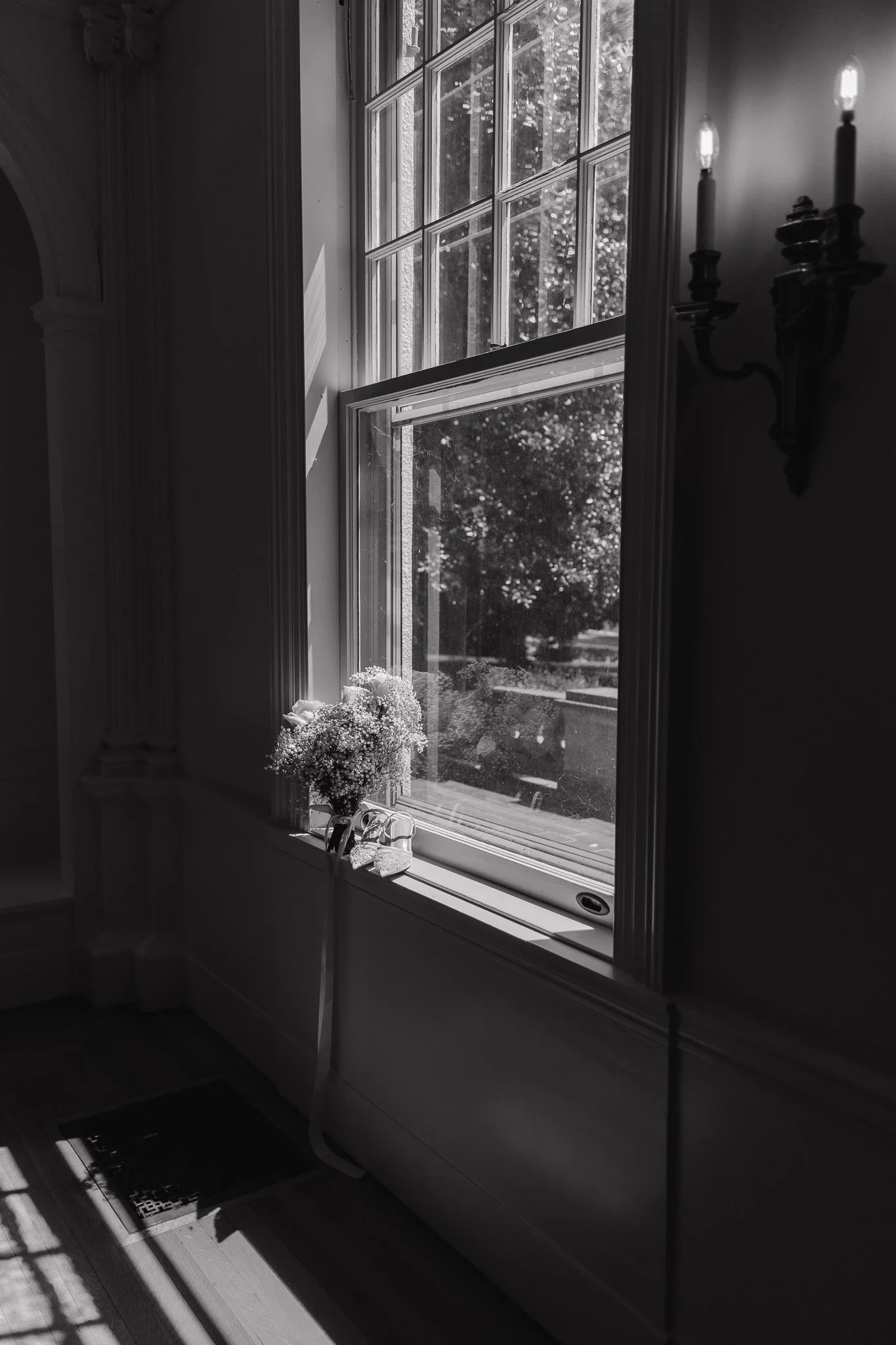 A black and white photo of a windowsill with a flower bouquet, a ribbon, and a small object, adjacent to a wall with a lit wall sconce light fixture.