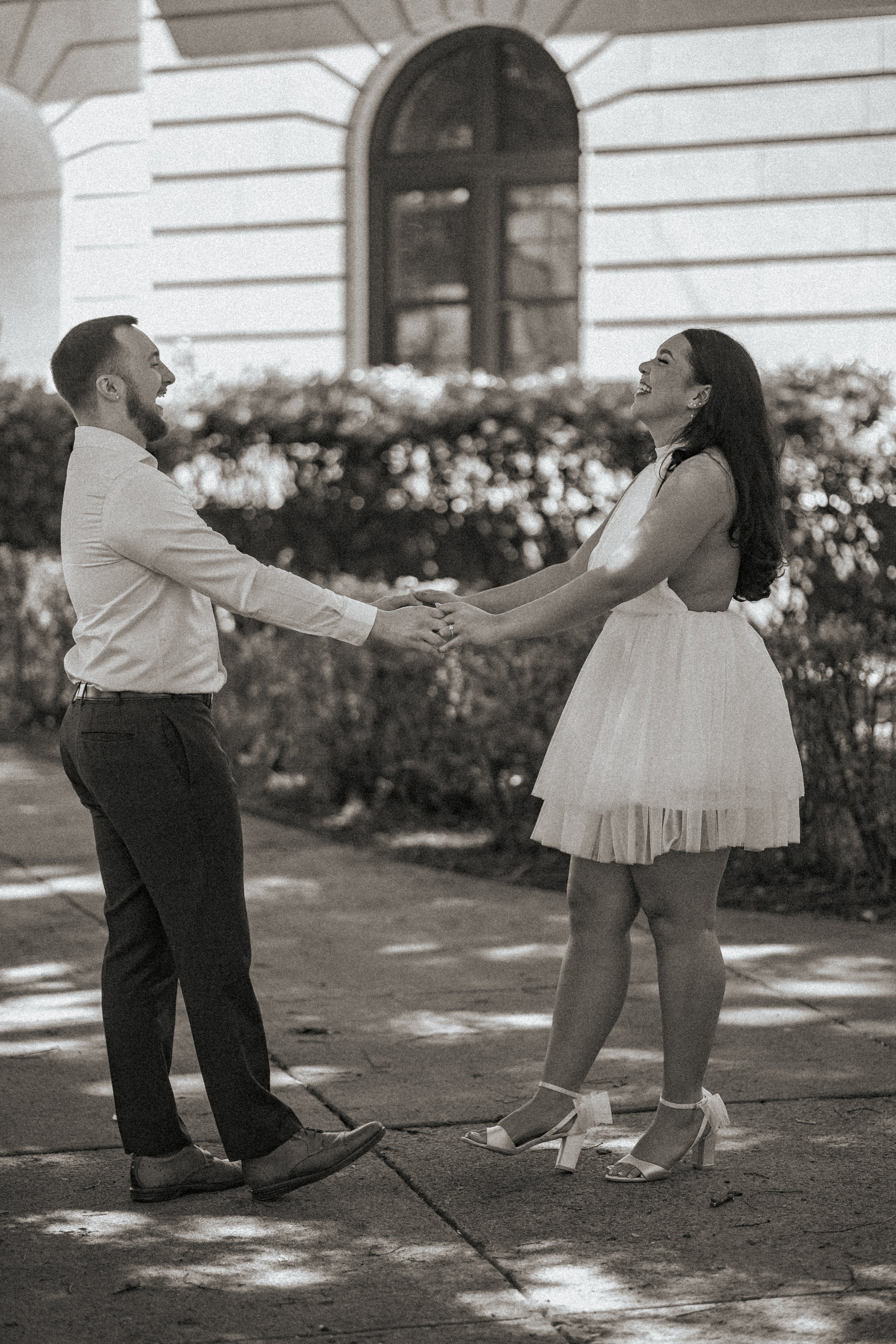 A couple holding hands and smiling outdoors, with the man wearing dress pants and a shirt, and the woman in a white dress and heels, standing on a sidewalk in front of a building with shrubs.