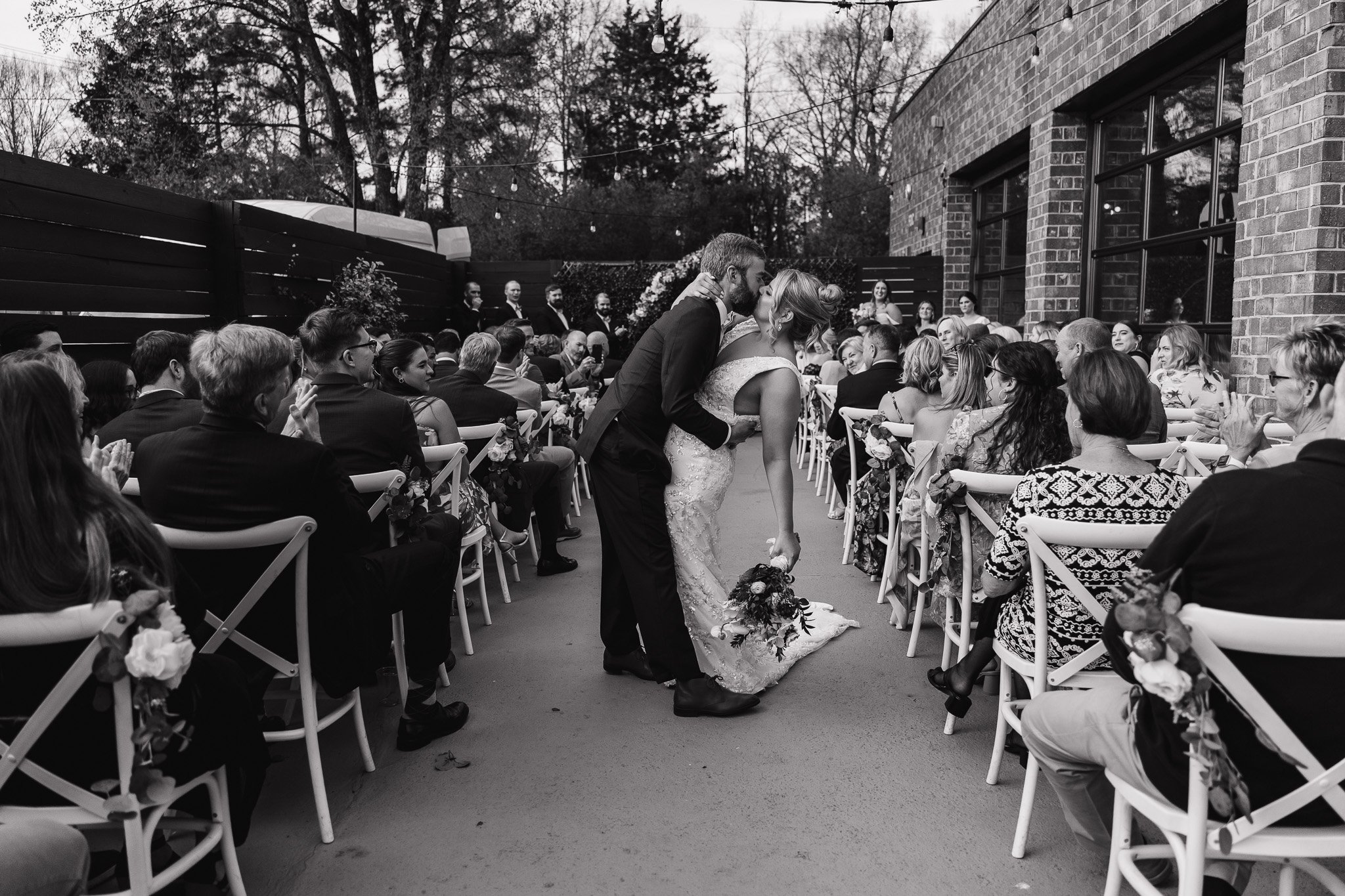 Bride and groom kissing in an aisle with guests seated on both sides during an outdoor wedding ceremony.