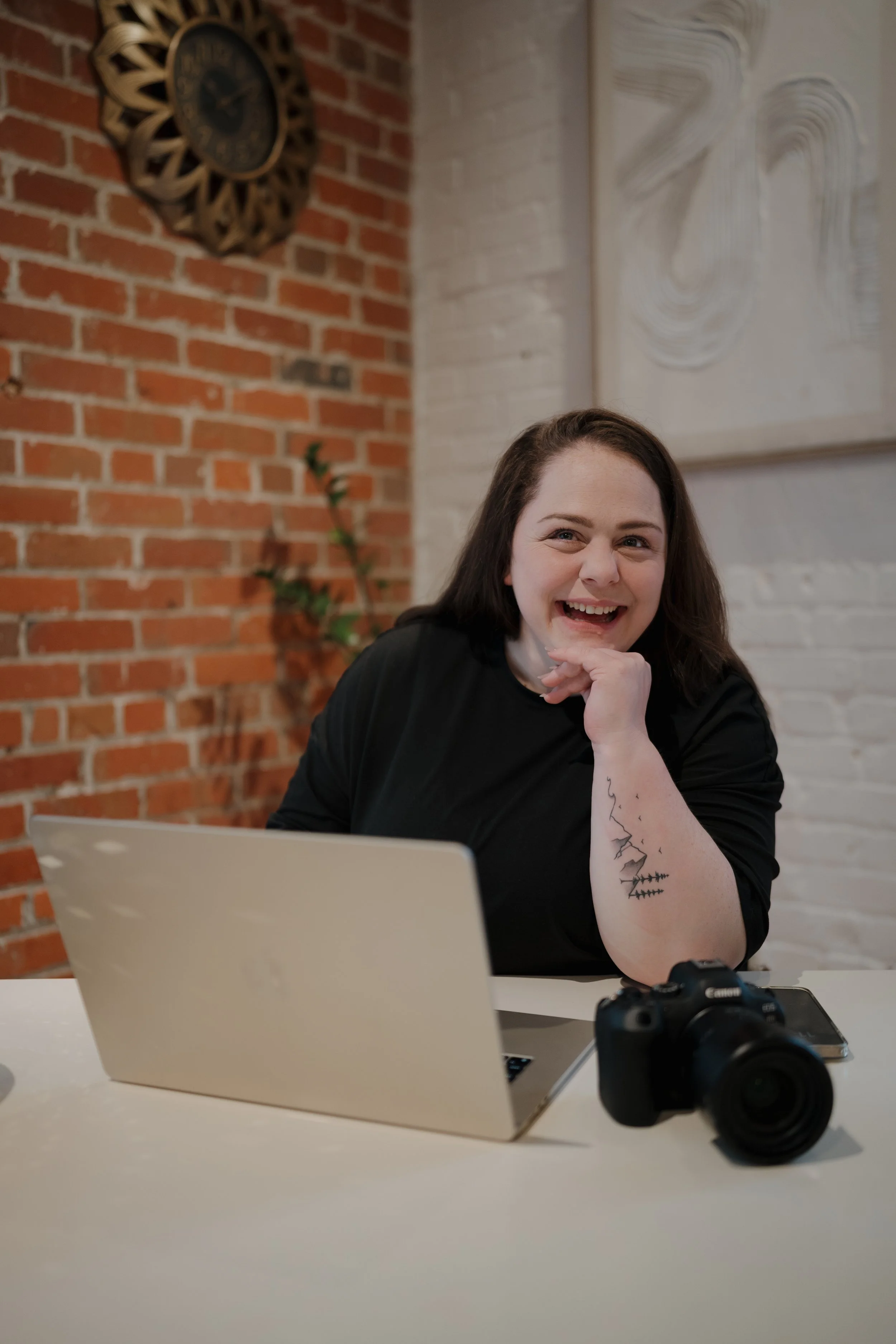 Person smiling at a desk with a laptop and camera, brick wall background.
