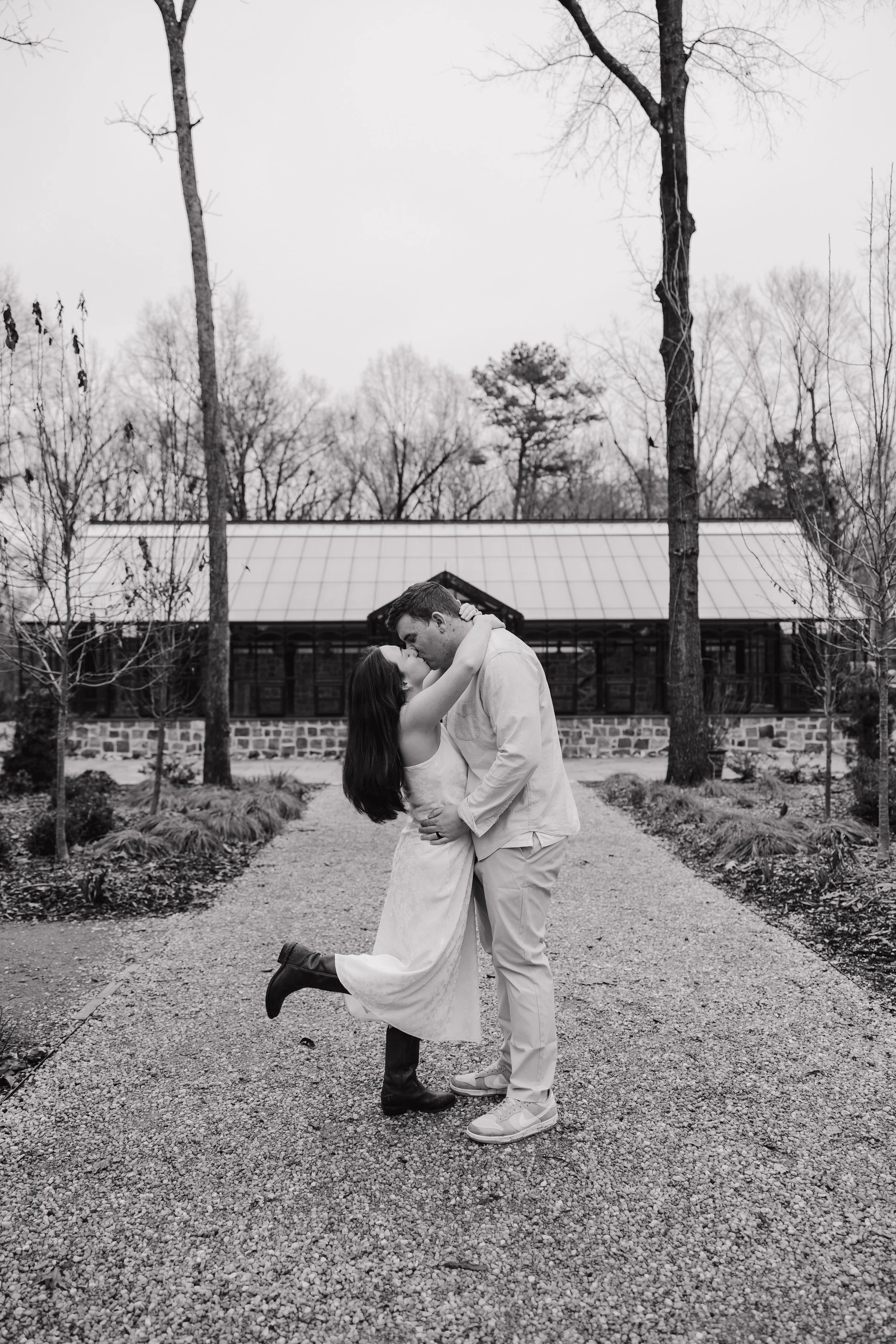 A couple sharing a kiss outdoors on a gravel path surrounded by trees, with a building in the background.