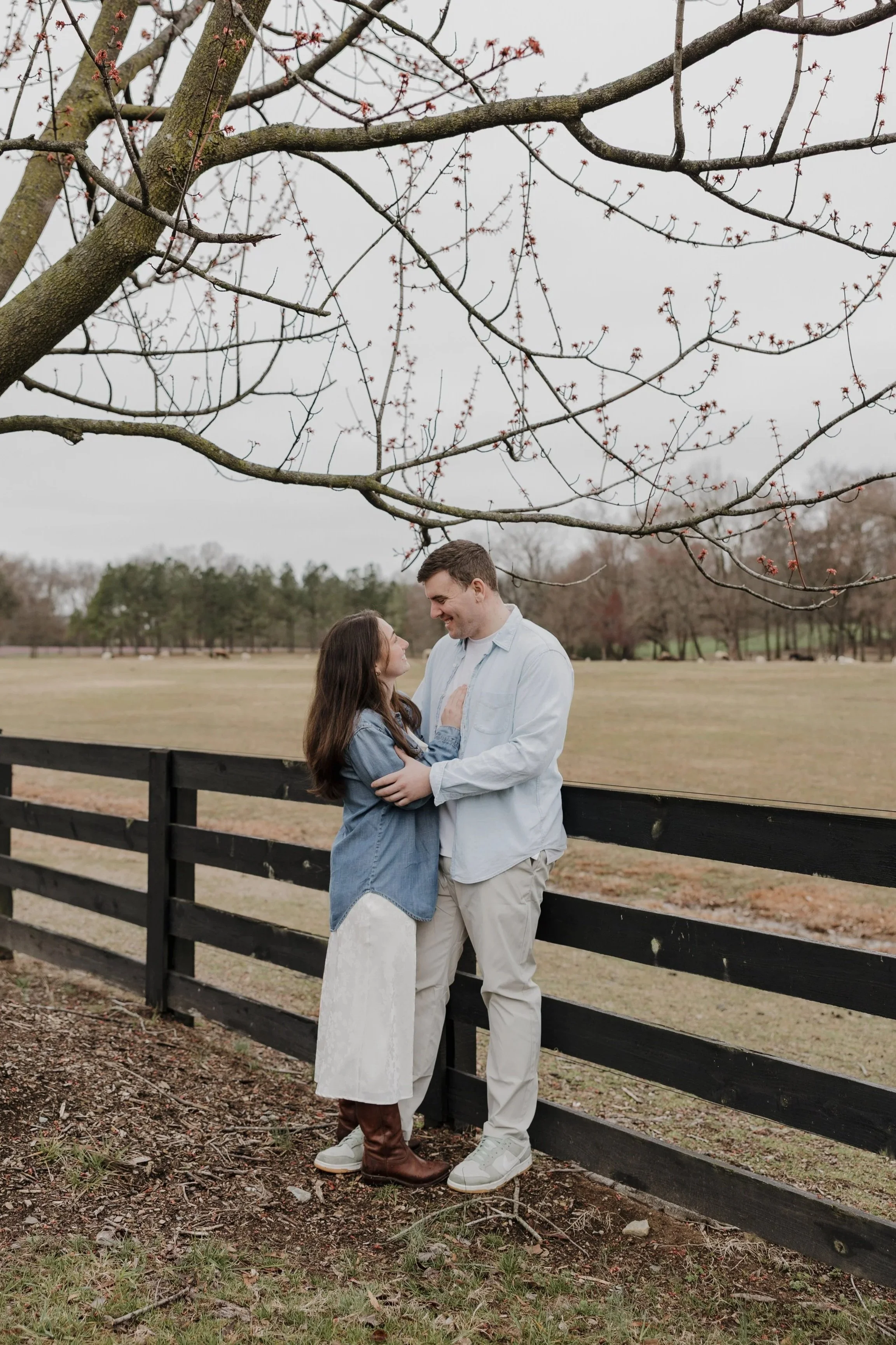 A couple standing close together near a black wooden fence, smiling at each other in a park with a large tree overhead and a field in the background.