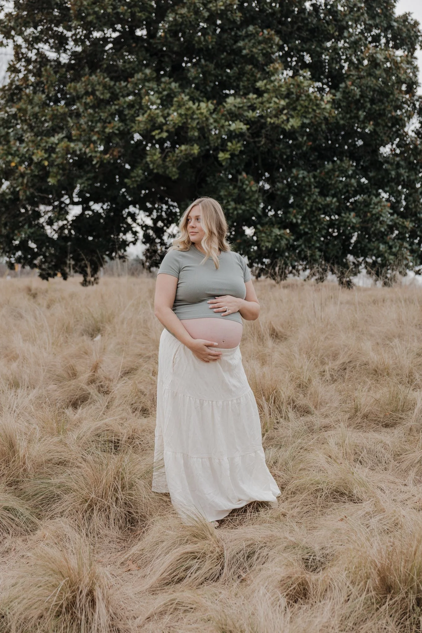  Dreamy maternity photos taken at NCMA Park in Raleigh, featuring a mother gently holding her belly in a quiet field setting 