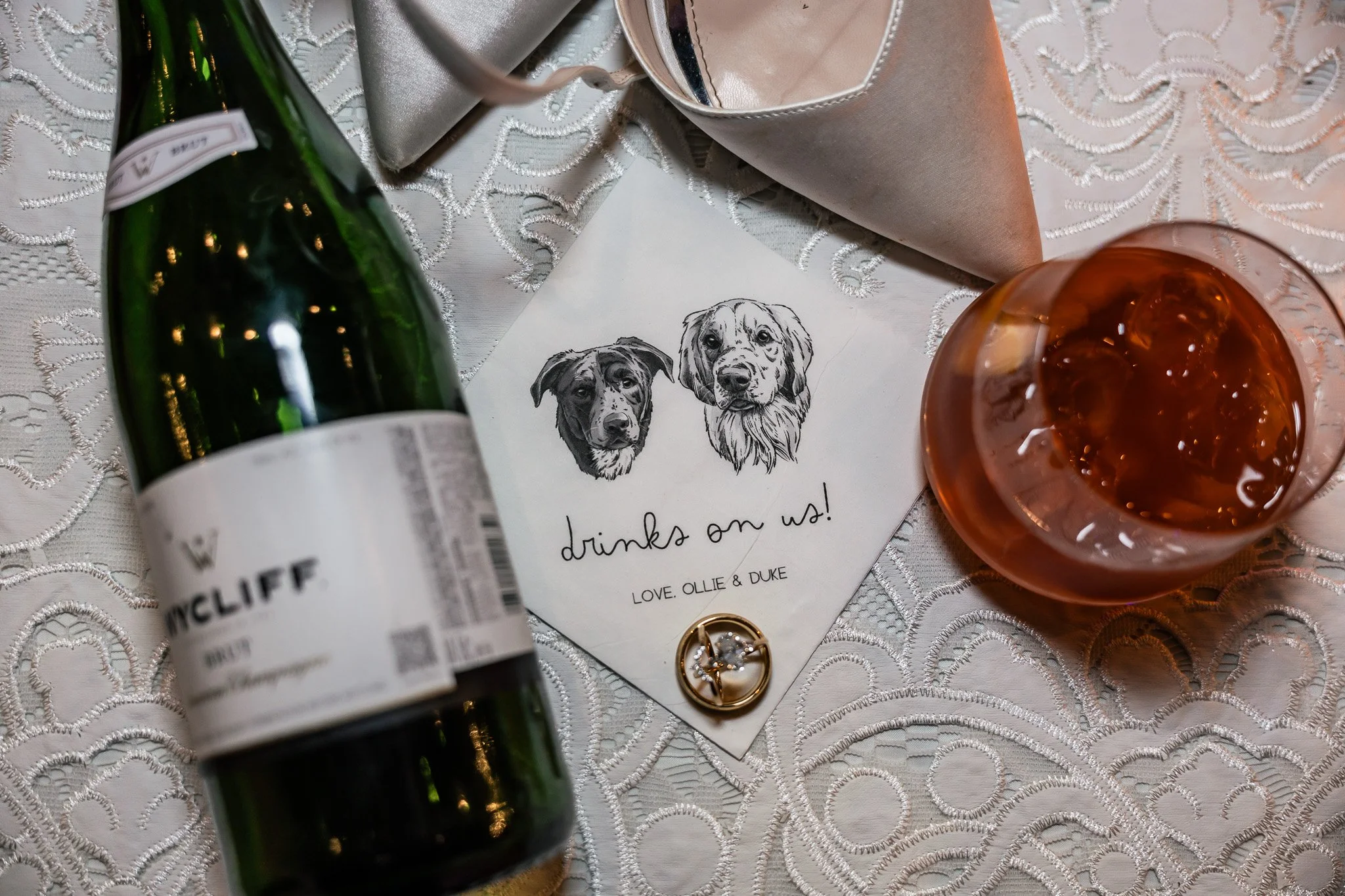 Champagne bottle, napkin with dog illustrations, rings, and cocktail on lace tablecloth.