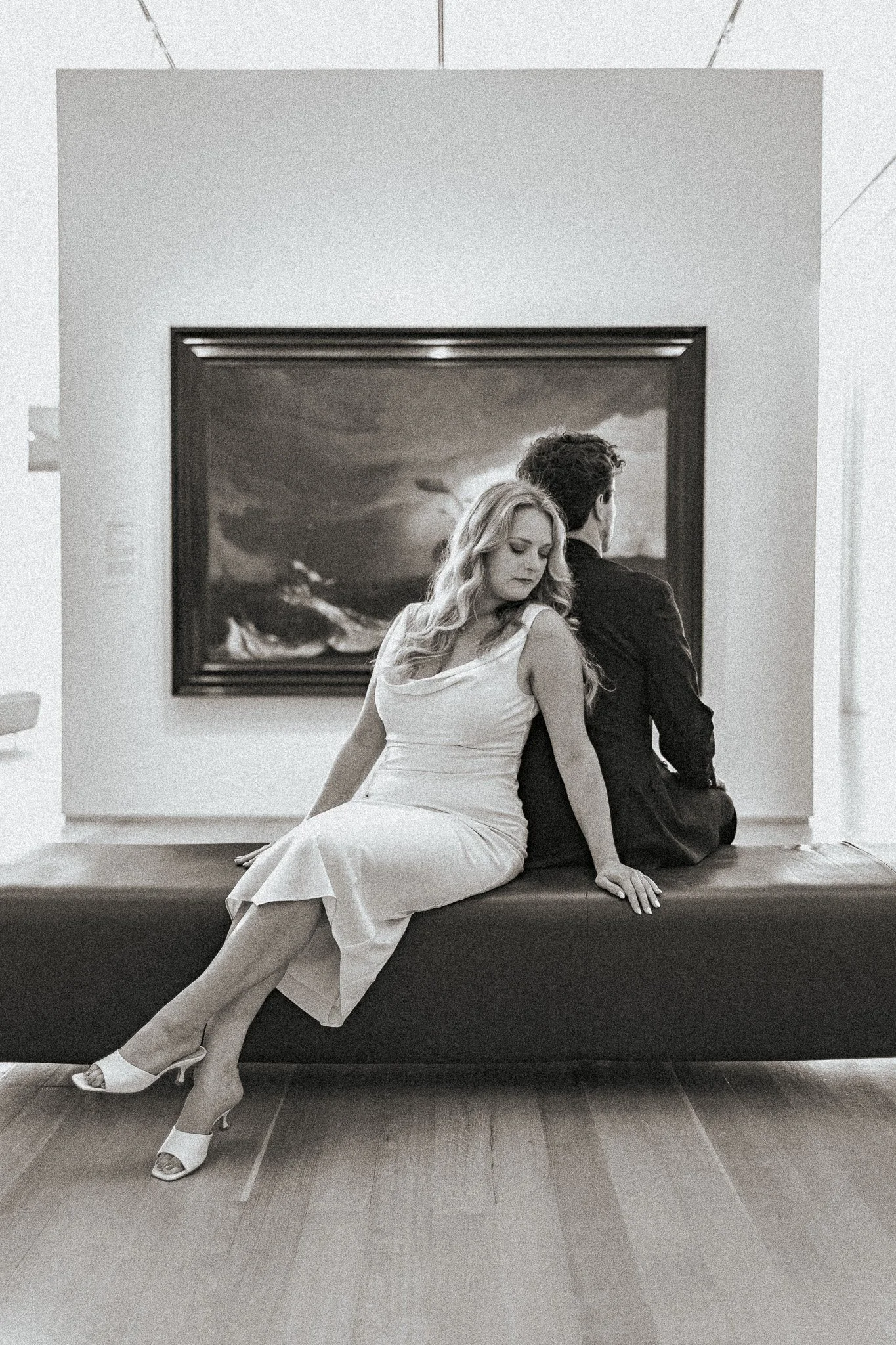 A black and white photo of a man and woman sitting back-to-back on a bench in an art gallery, with a large painting in the background.