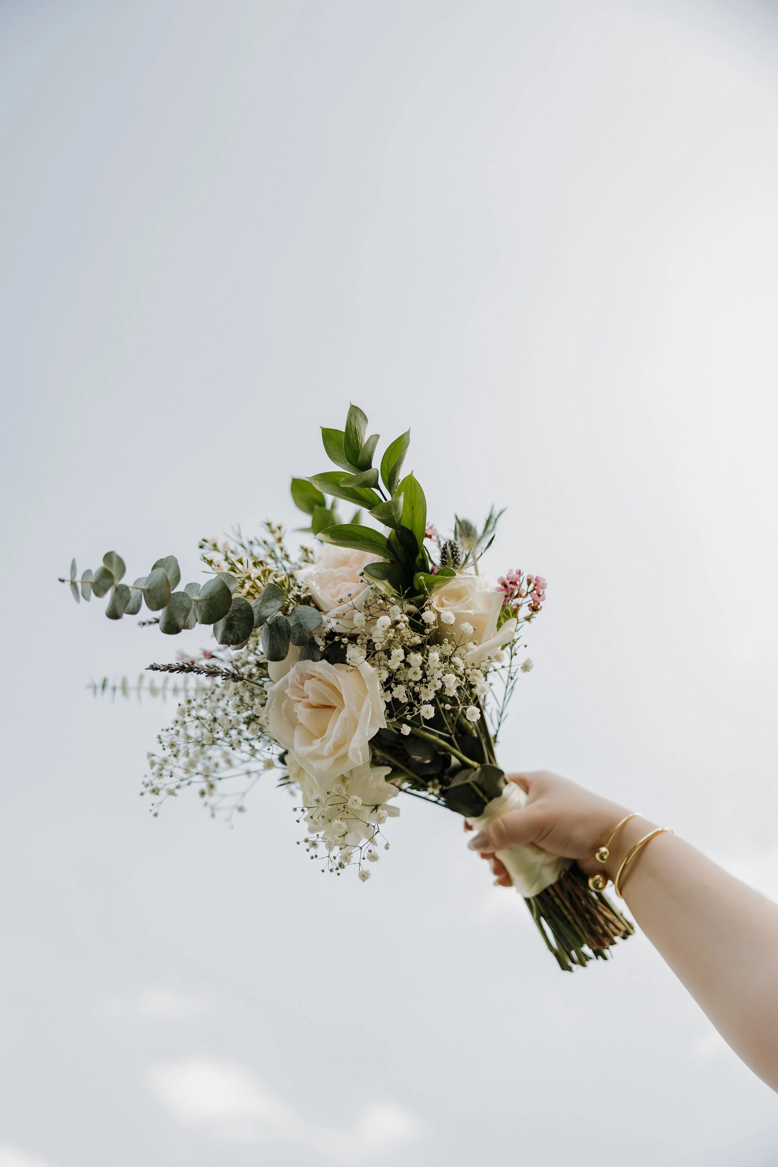 Person holding a bouquet of white roses and green foliage against a cloudy sky.