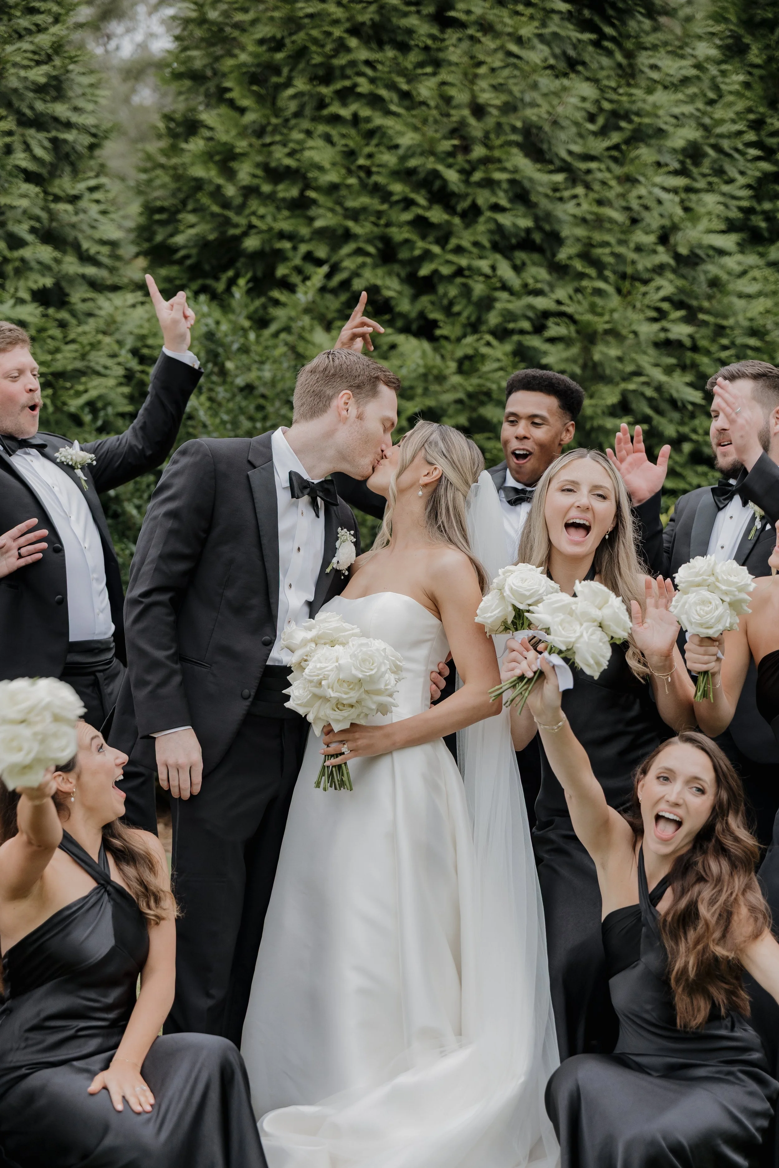 A newlywed couple kissing, surrounded by wedding party members celebrating outdoors with greenery in the background.