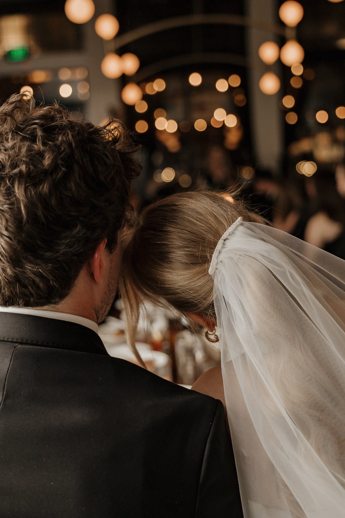Bride and groom from behind, bride wearing veil, in dimly lit room with bokeh lights.